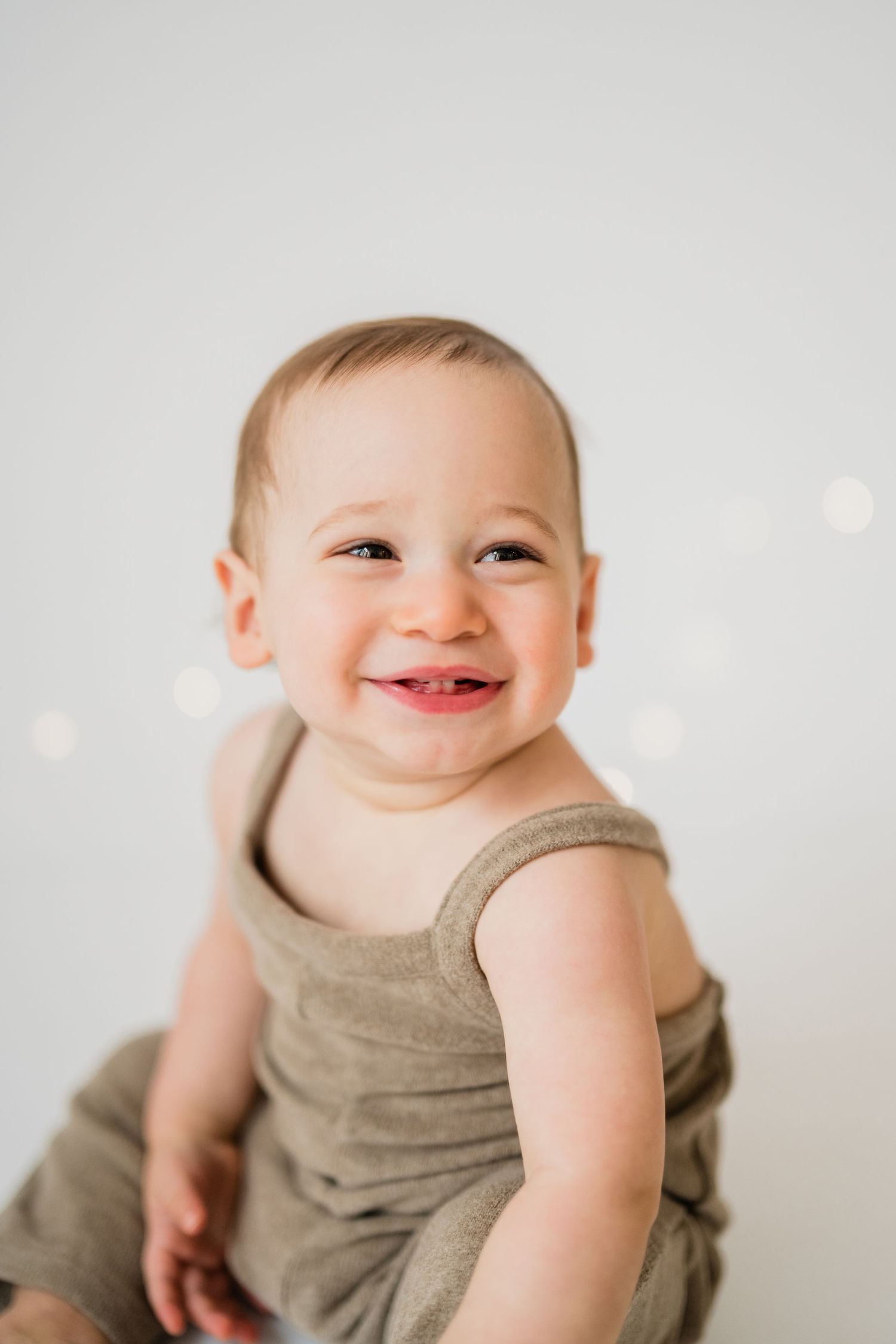 Smiling baby in olive romper sits against white background with soft bokeh lights, showing first teeth.