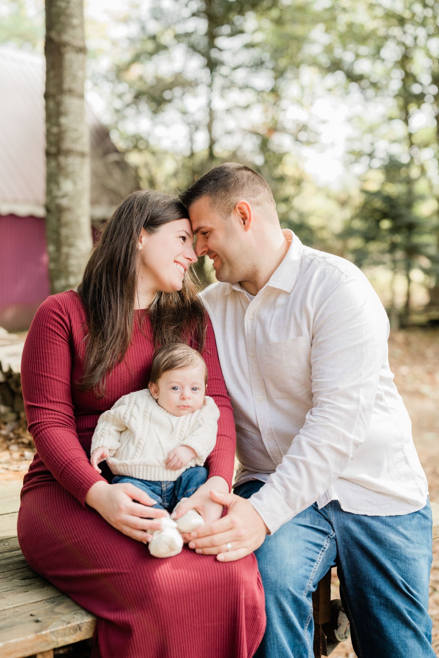 A young couple tenderly touch foreheads while holding their baby in a wooded outdoor family portrait session.