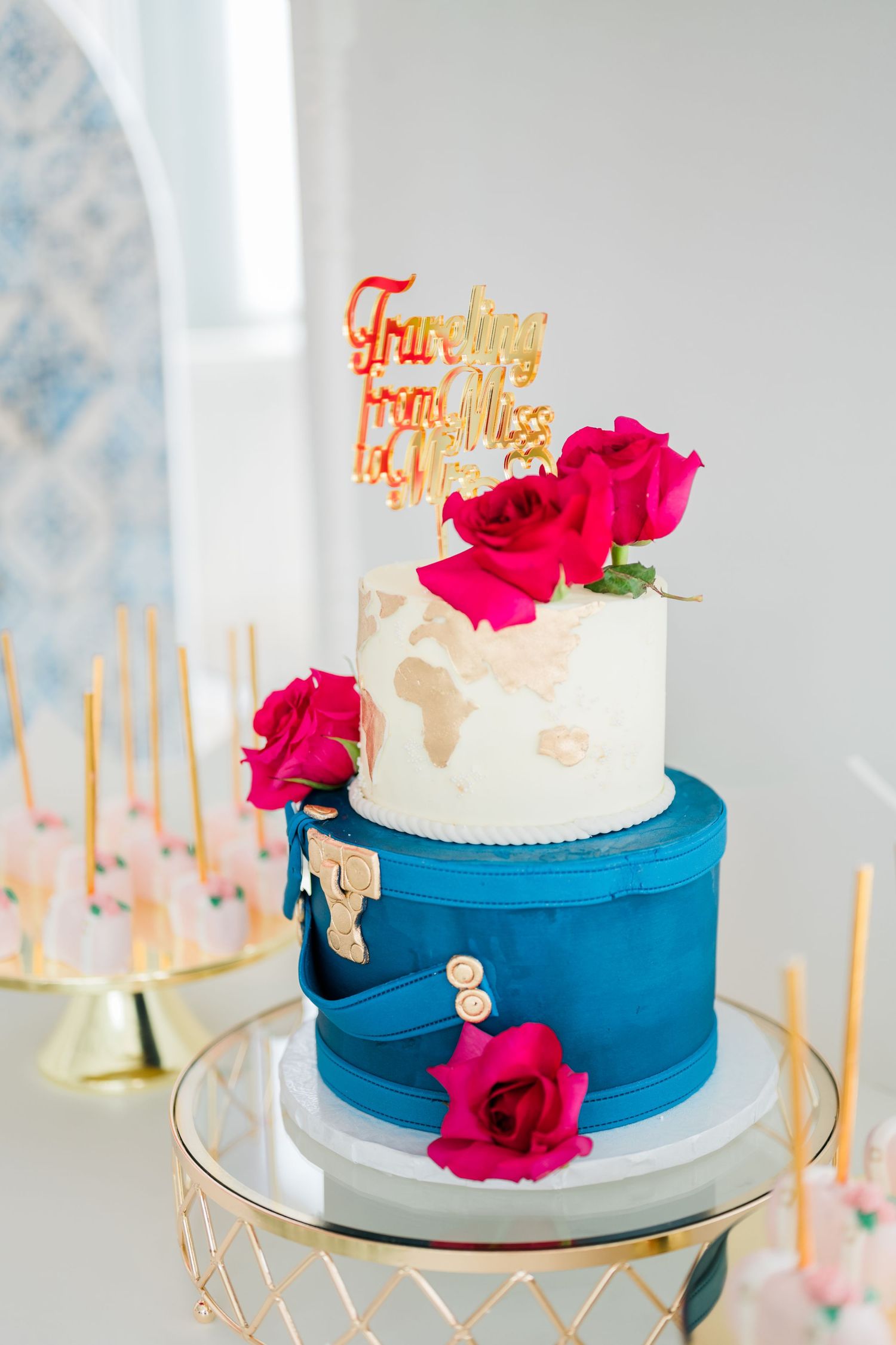 Two-tiered wedding cake with blue base and pink flowers topped with gold text decorations.