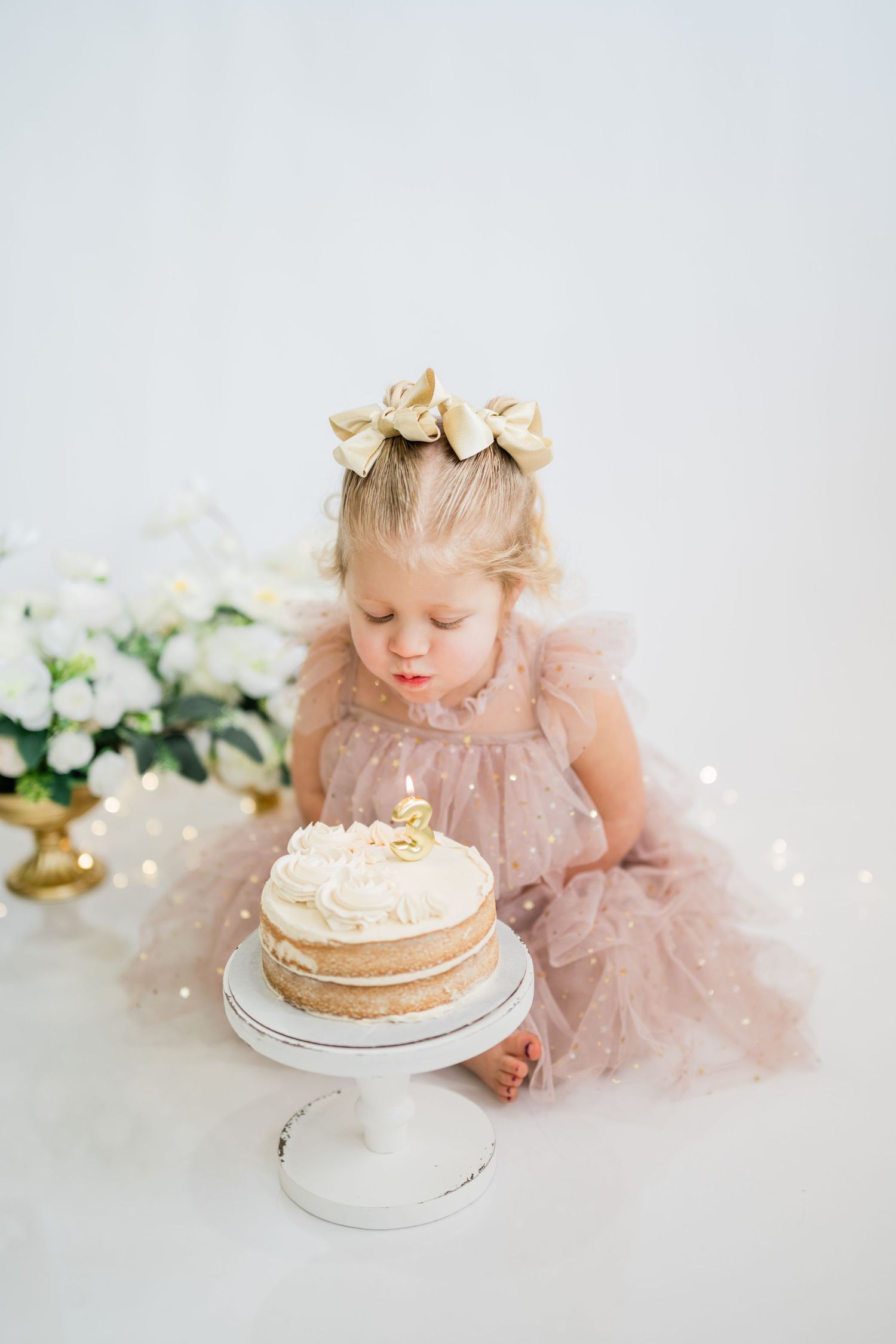 Toddler girl in pink tutu dress with gold bow blowing out candle on birthday cake on white cake stand.