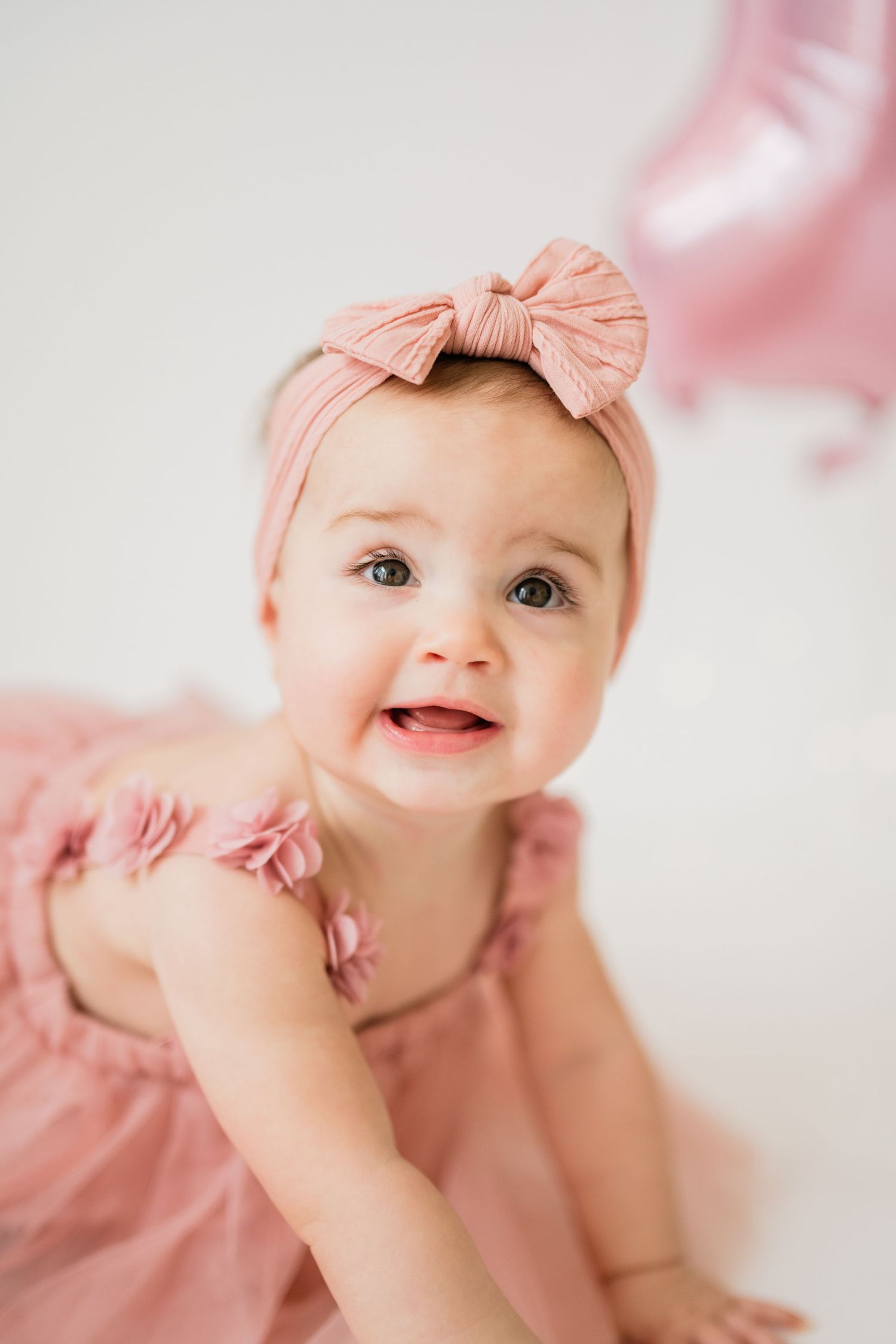 Adorable baby girl in pink dress and bow headband smiling, with pink balloon in background.