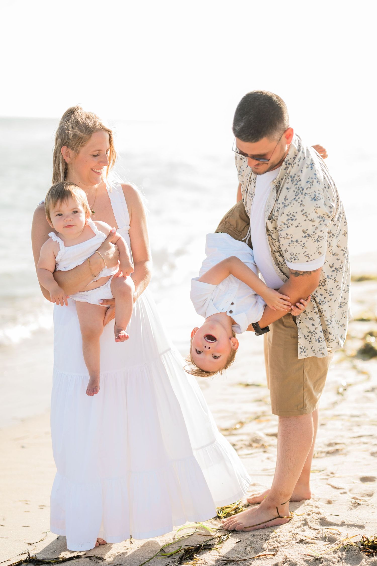 Happy family enjoying a sunny beach day, parents playfully holding two young children on sandy shore.