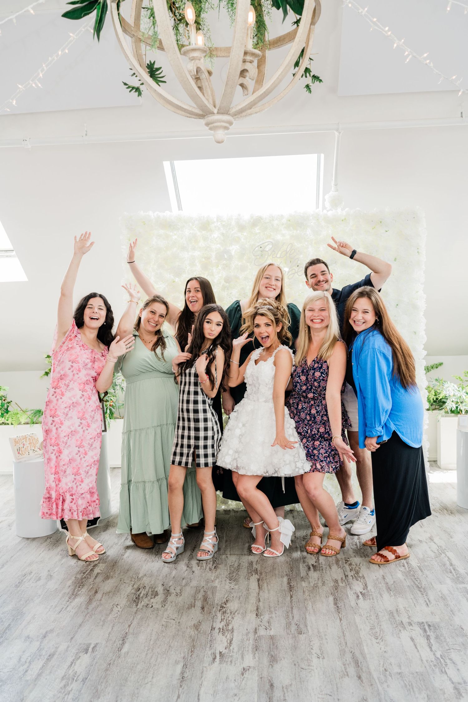 A group of friends pose joyfully against a white floral wall in summer dresses.