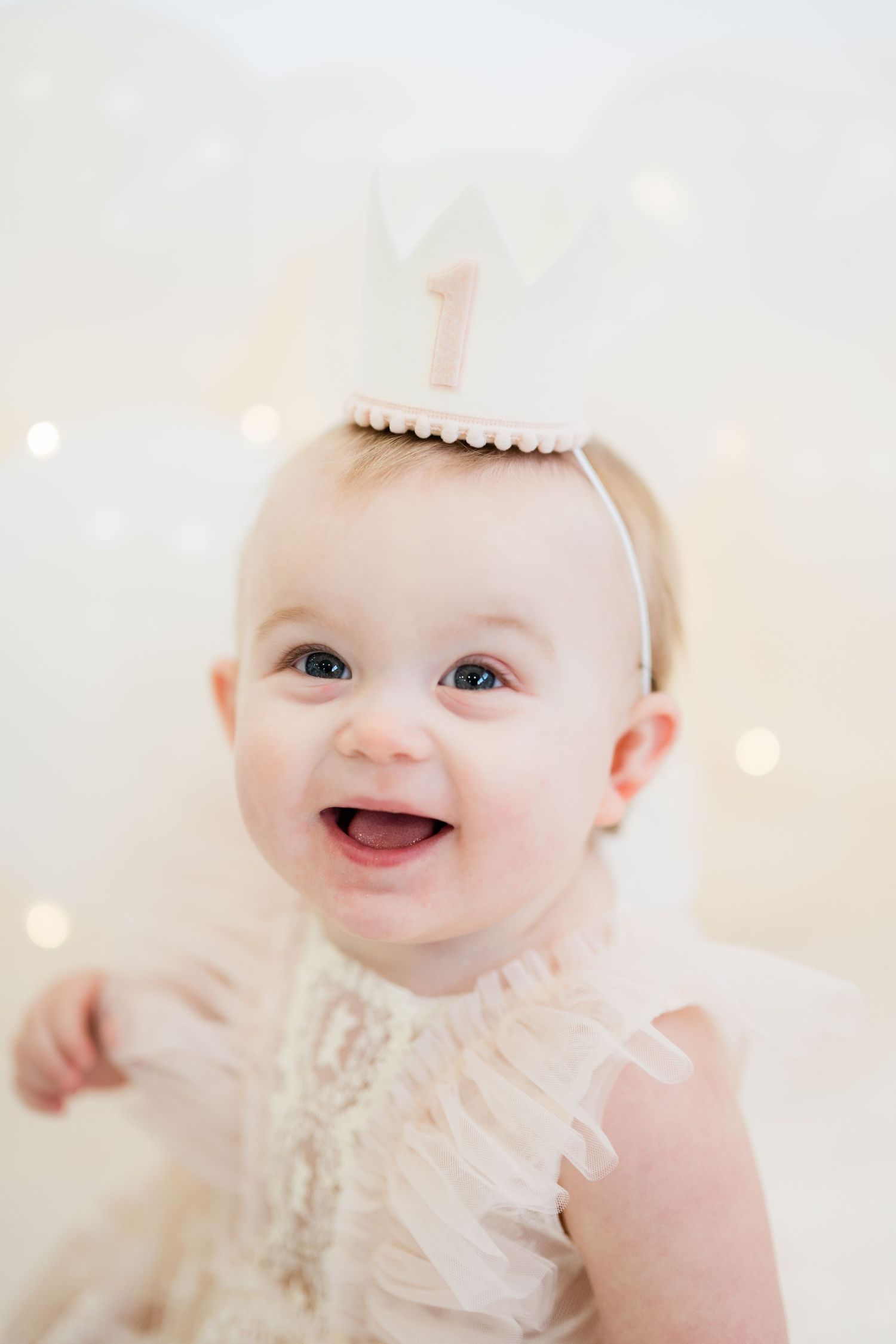 Smiling baby girl wearing a white number 1 birthday crown headband, celebrating her first birthday in a soft white setting.
