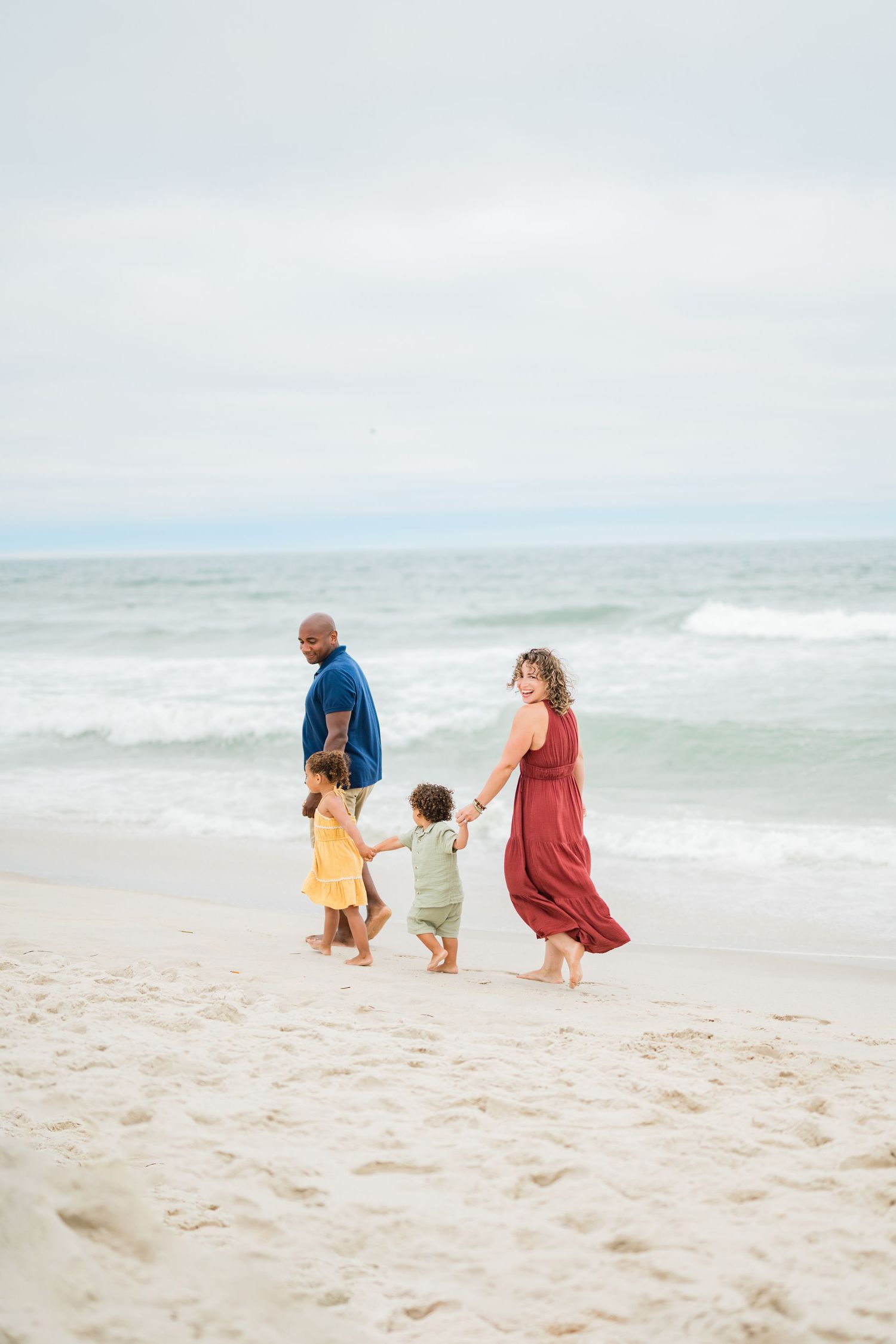 Family of four walking hand-in-hand on a sandy beach, parents in red and blue with two young children in yellow and green.