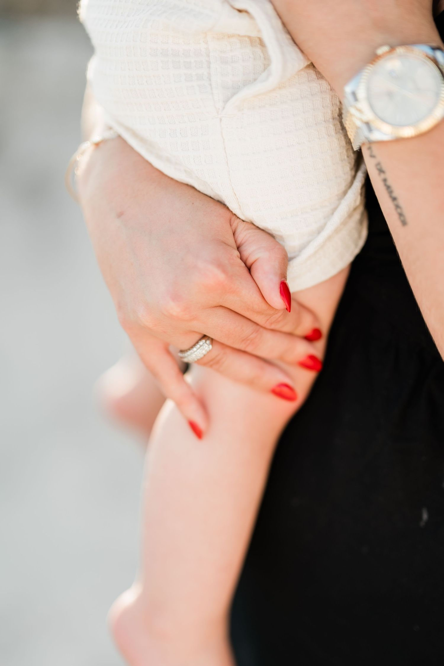 Close-up of a woman's hand with red nails and diamond ring, wearing a gold watch, holding a baby.