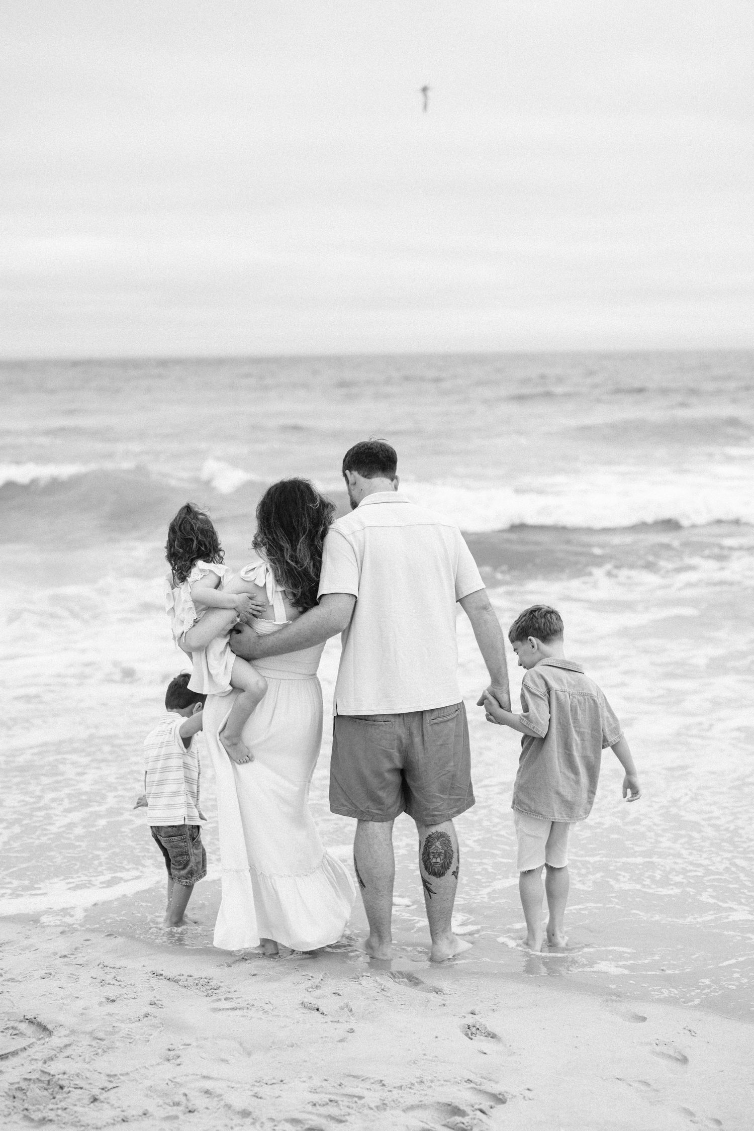 Black and white photo of a family of five standing together at the ocean's edge, facing the waves on a beach.