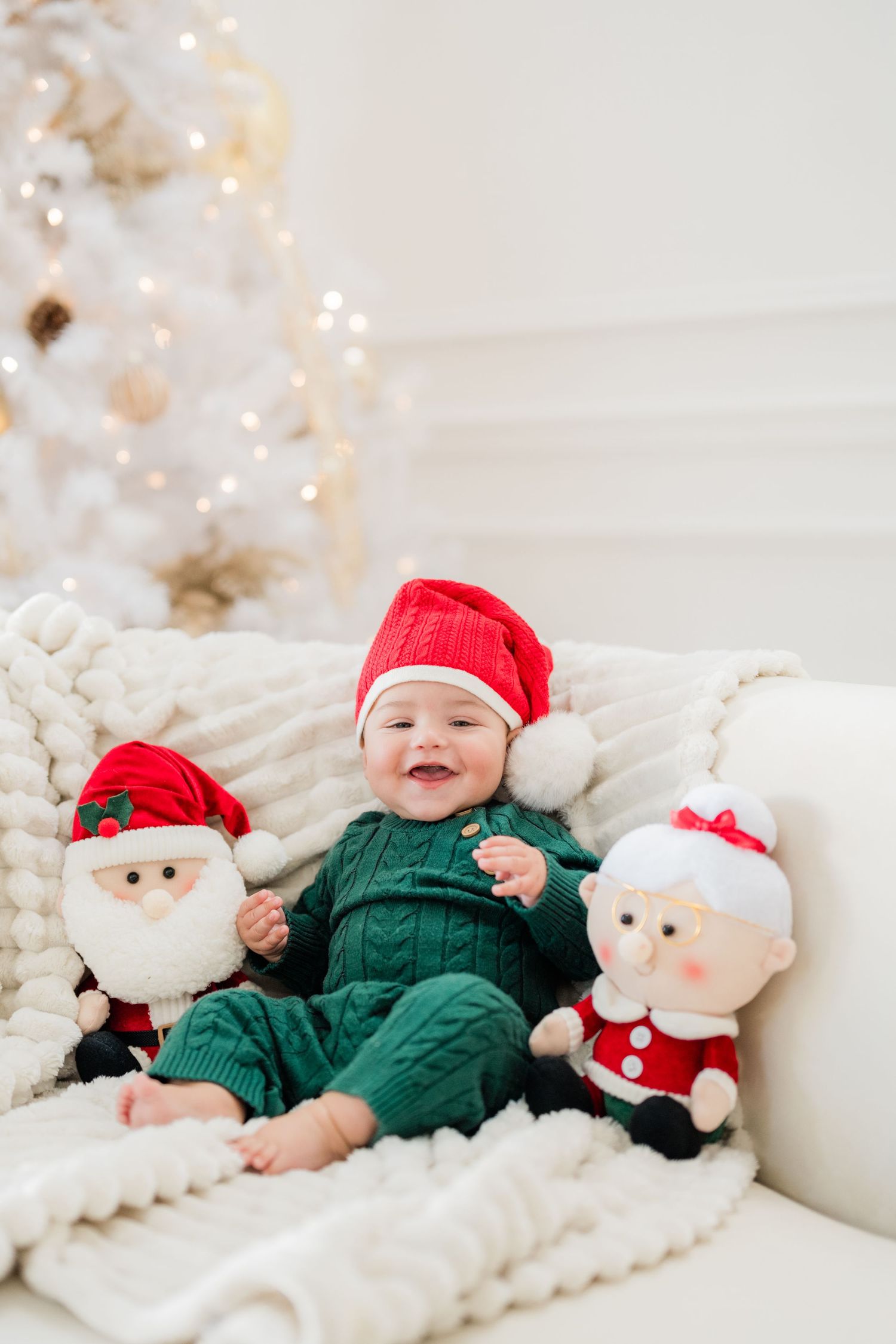 Baby in green knit outfit and Santa hat smiles between Santa and Mrs. Claus plush toys near white Christmas tree.