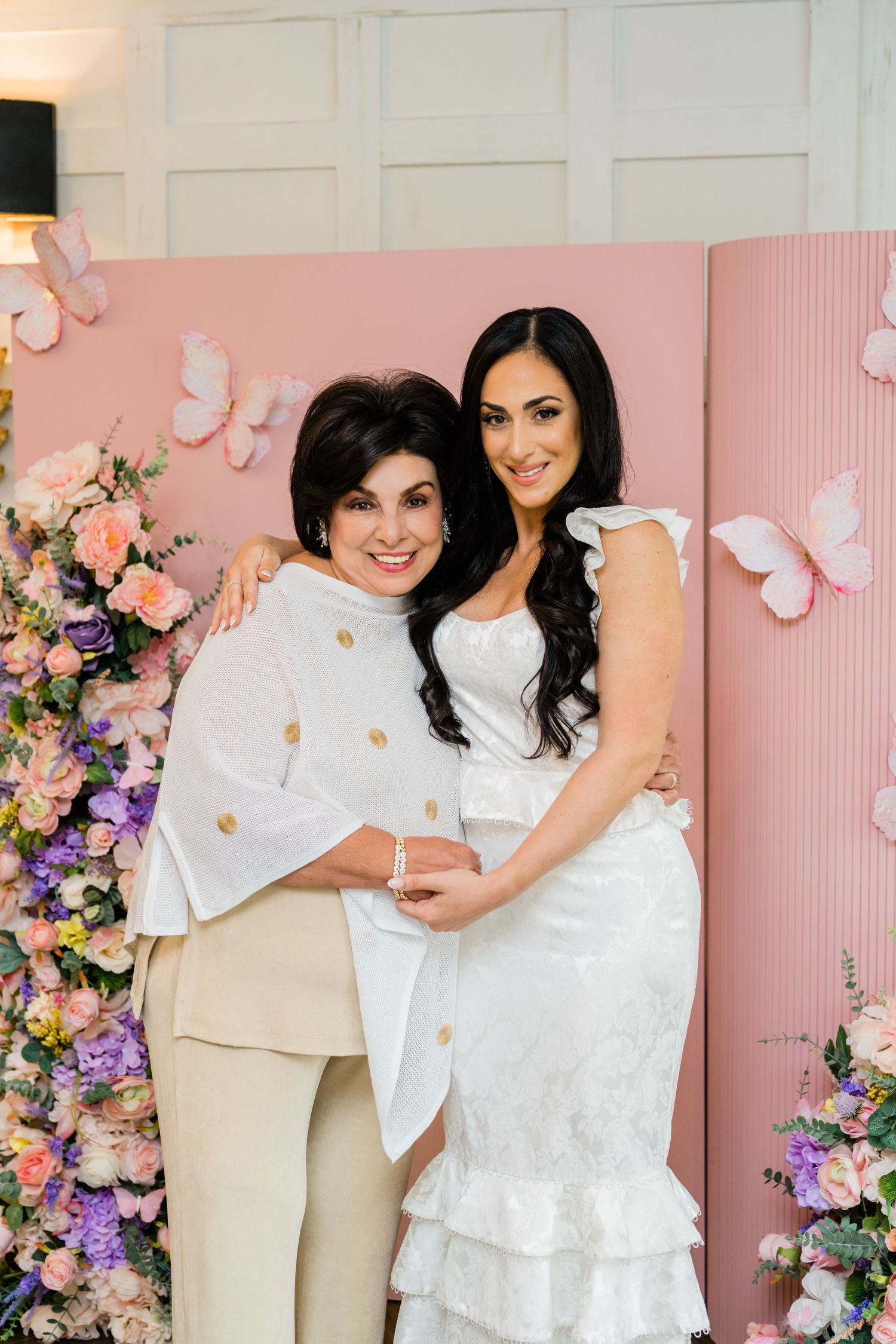 Two people pose together against a pink backdrop decorated with butterflies and flowers.