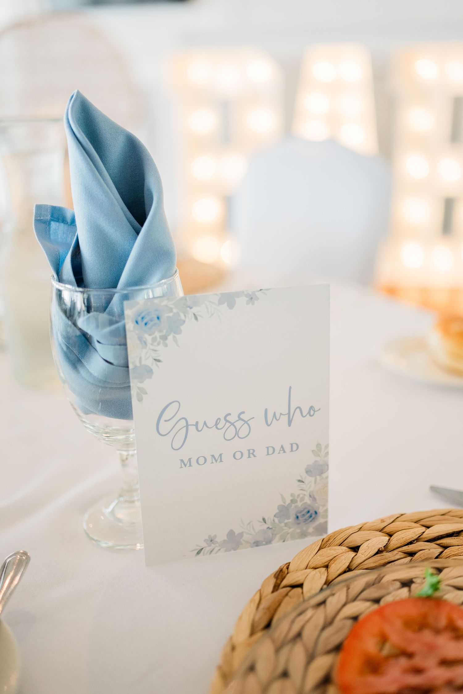 Elegant place setting with light blue napkin and 'Guess who Mom or Dad' card on white tablecloth.