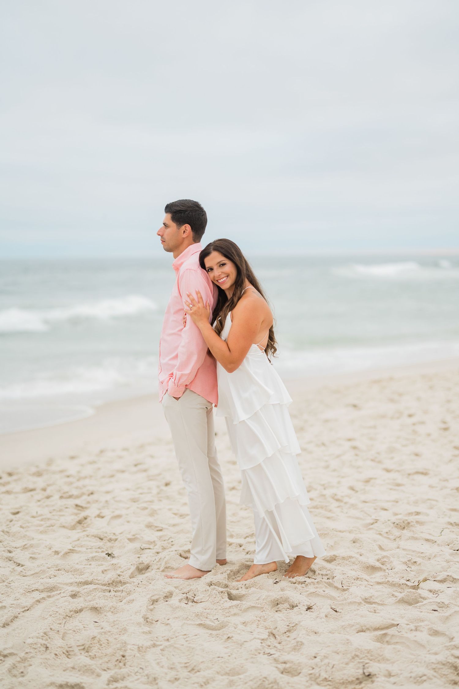 Couple posing on sandy beach, woman in white ruffled dress hugging man in pink shirt, ocean waves in background.
