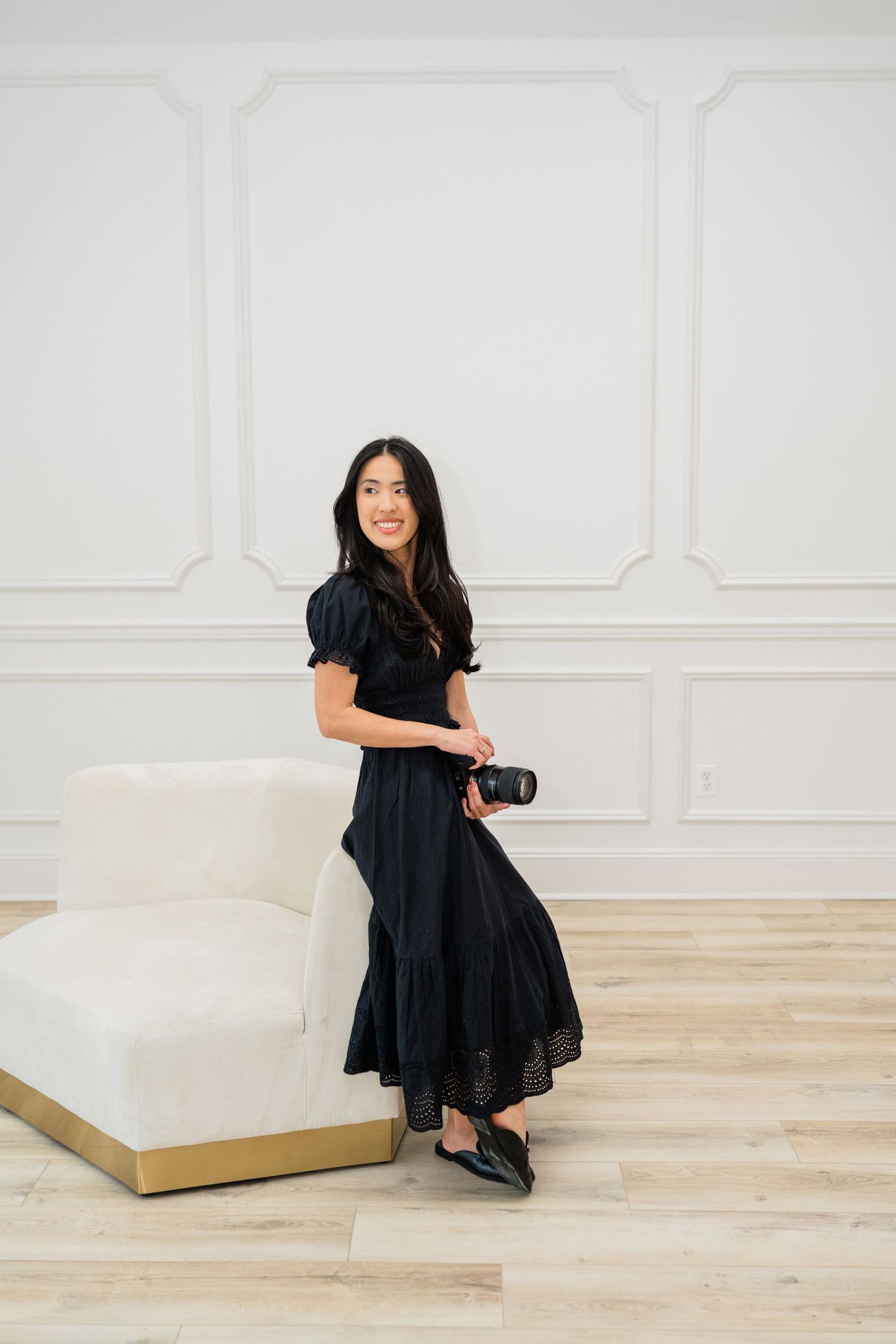 Person in black dress holding camera while posing near white couch.