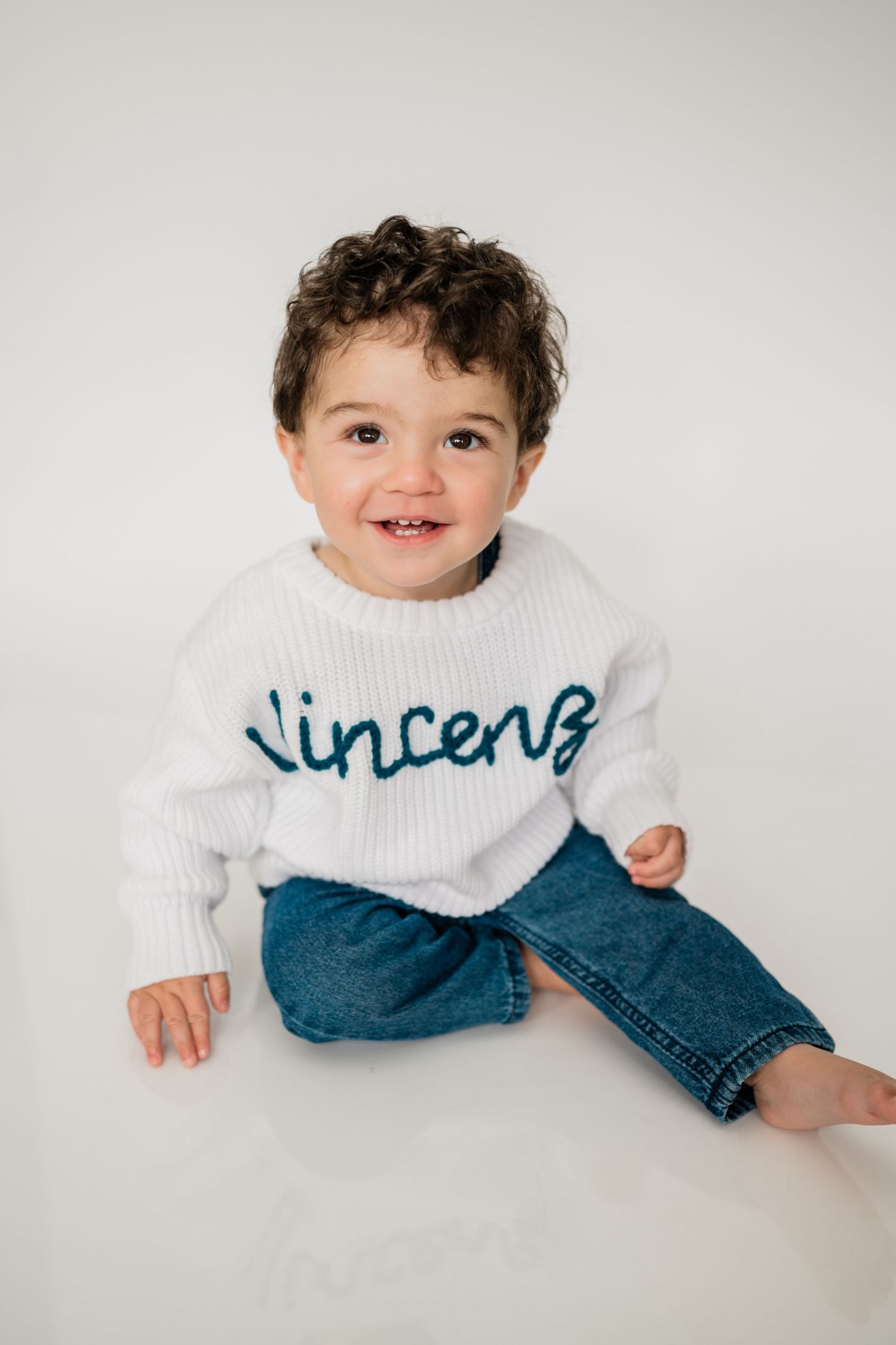Smiling toddler boy wearing a white knit sweater with name Vincent in teal, paired with blue jeans.
