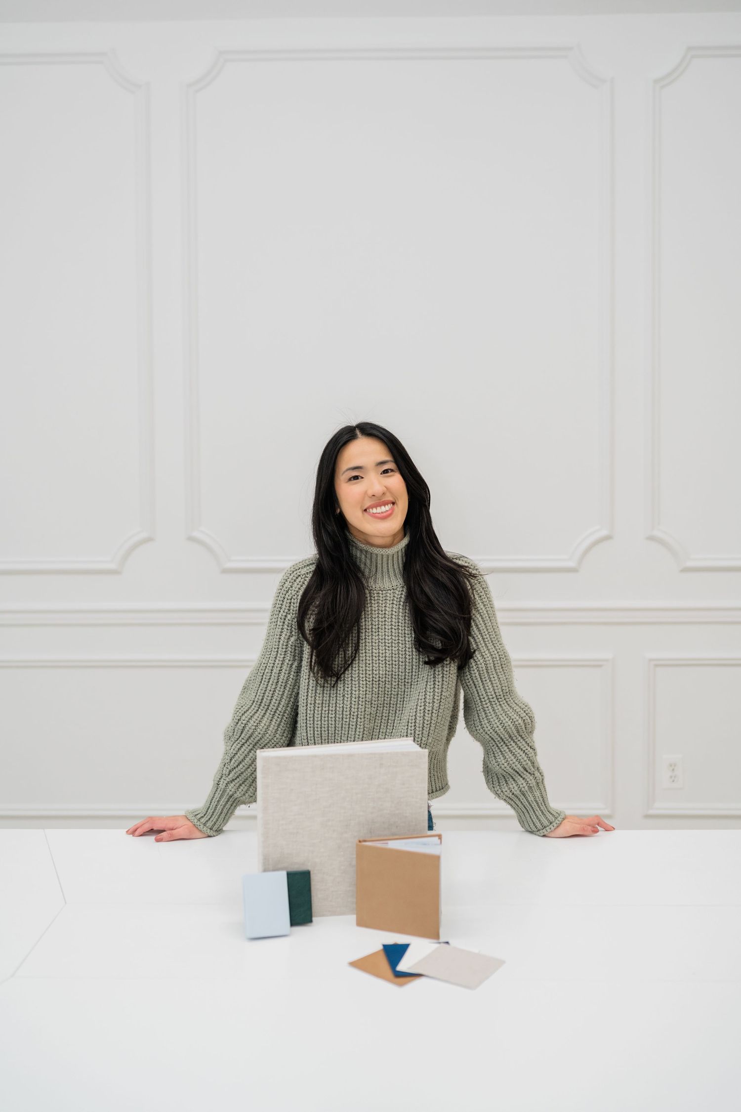 Person in sage green sweater sitting at white desk against white wall.