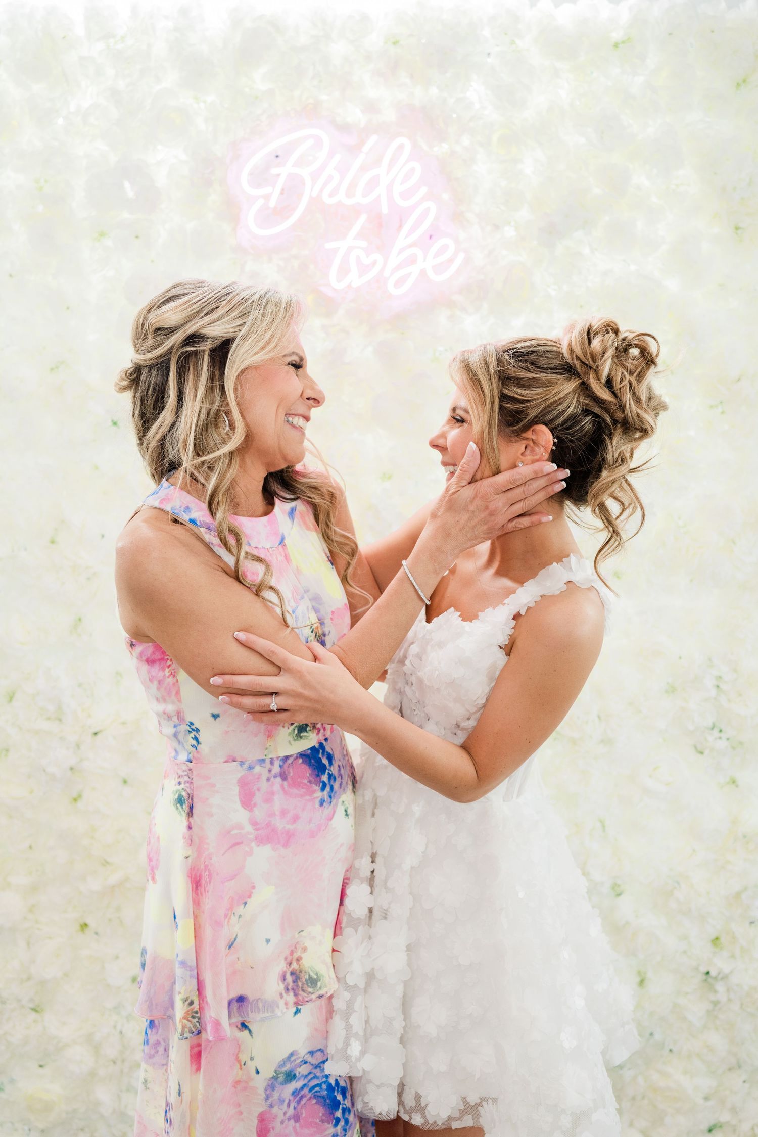 Two friends share an emotional moment against a white floral backdrop with pink neon lighting.