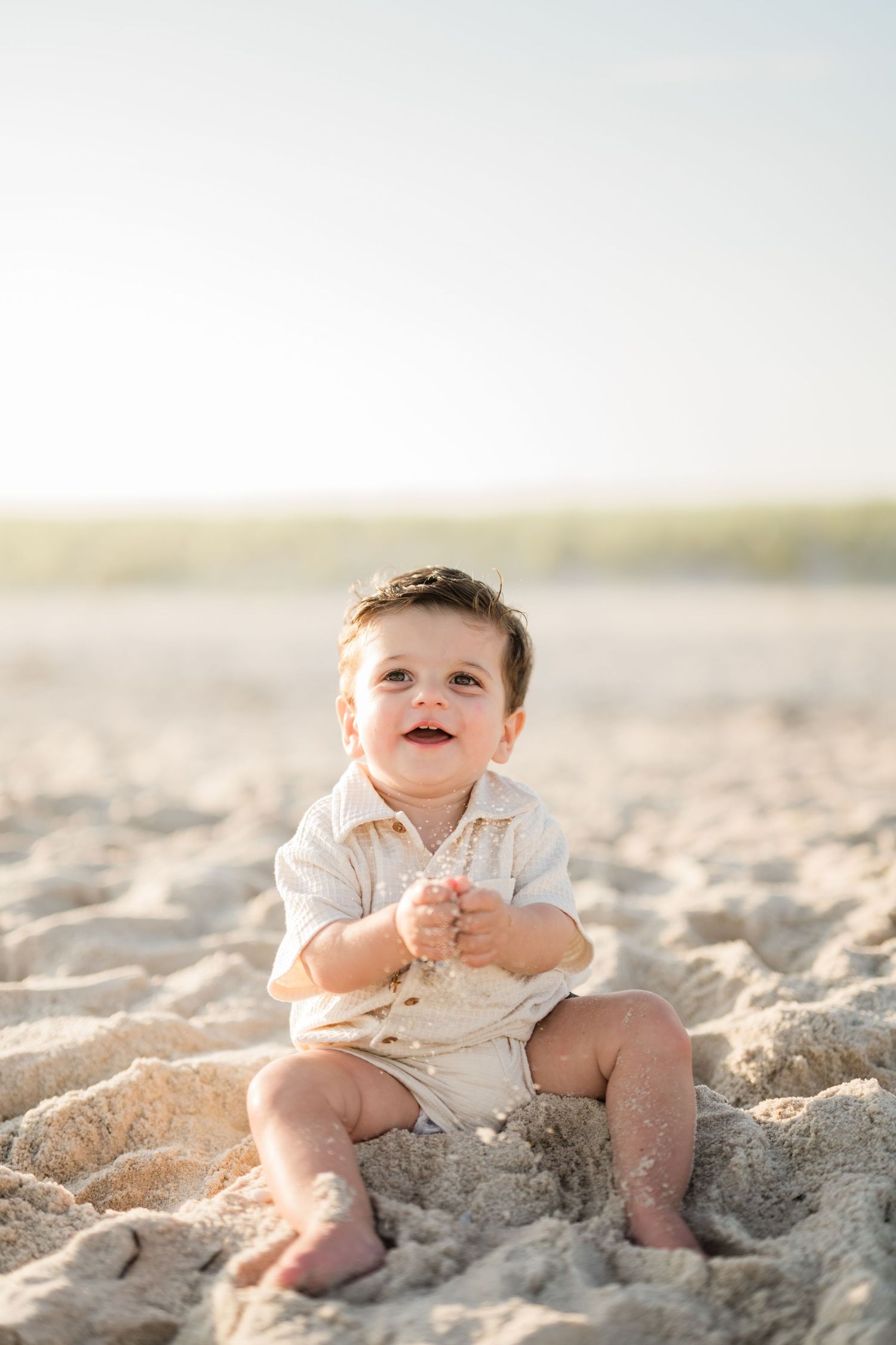 Happy baby sitting on sandy beach in white romper, smiling up at sky on a bright sunny day.