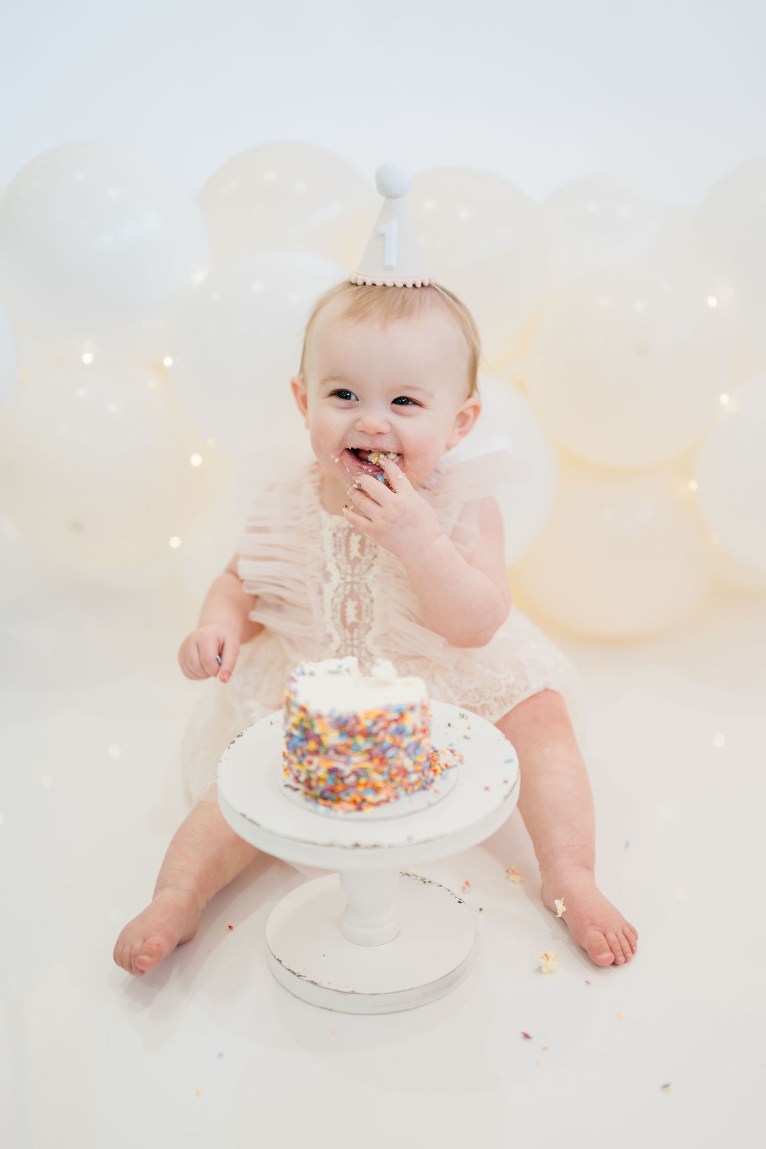 Baby girl in pink tutu and party hat smashes first birthday cake surrounded by white balloons.