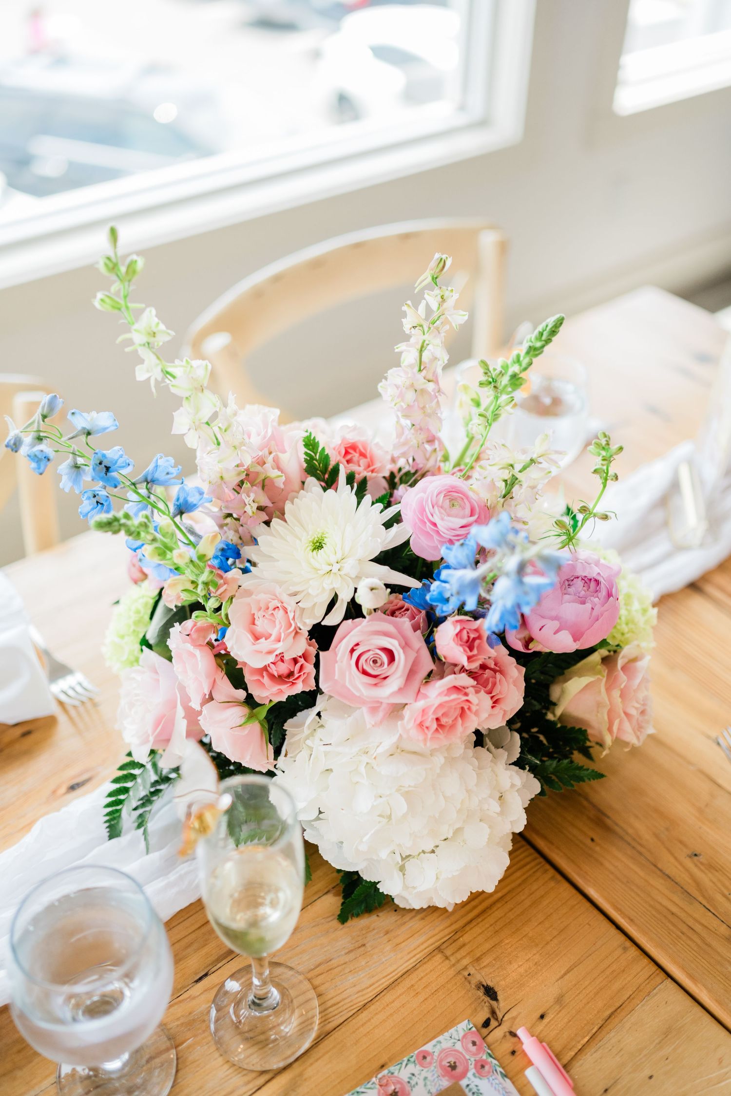 Elegant floral centerpiece with pink roses, white hydrangeas, and blue delphinium on a wooden table with wine glasses.