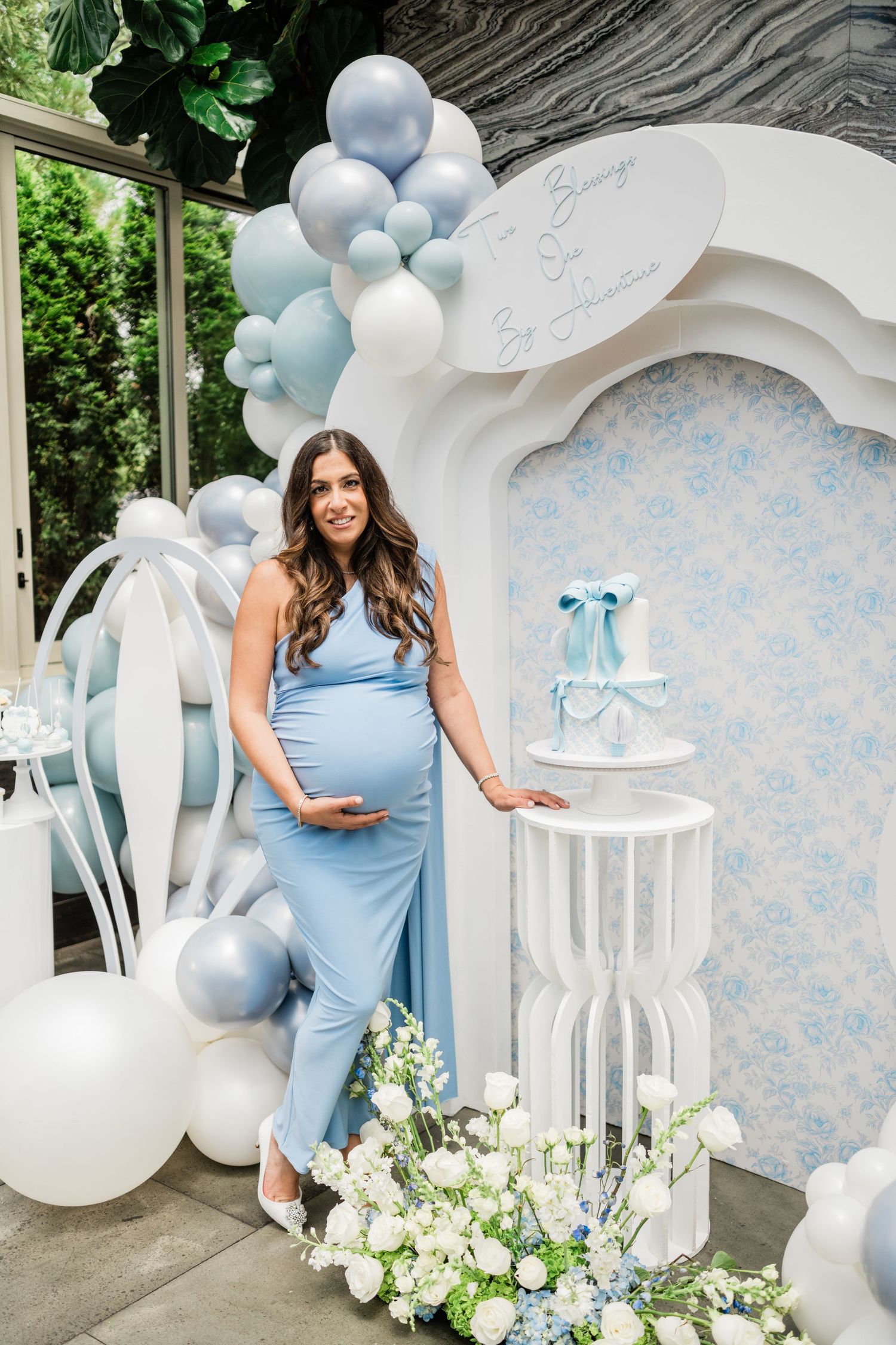 Baby shower setup with white arch backdrop, blue balloons, and white floral arrangements.