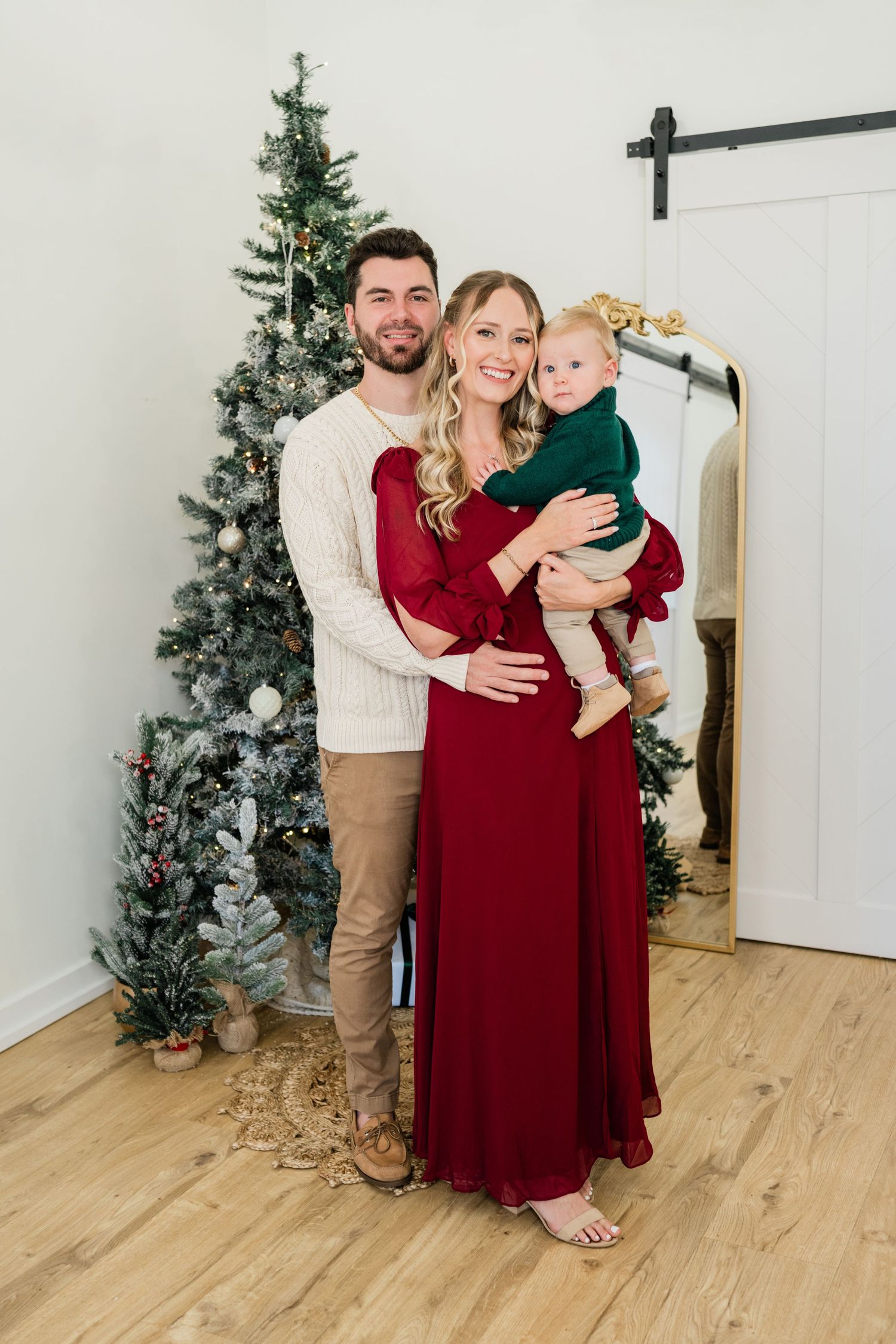 Family Christmas portrait with parents in red and cream holding baby in green dress by decorated tree.