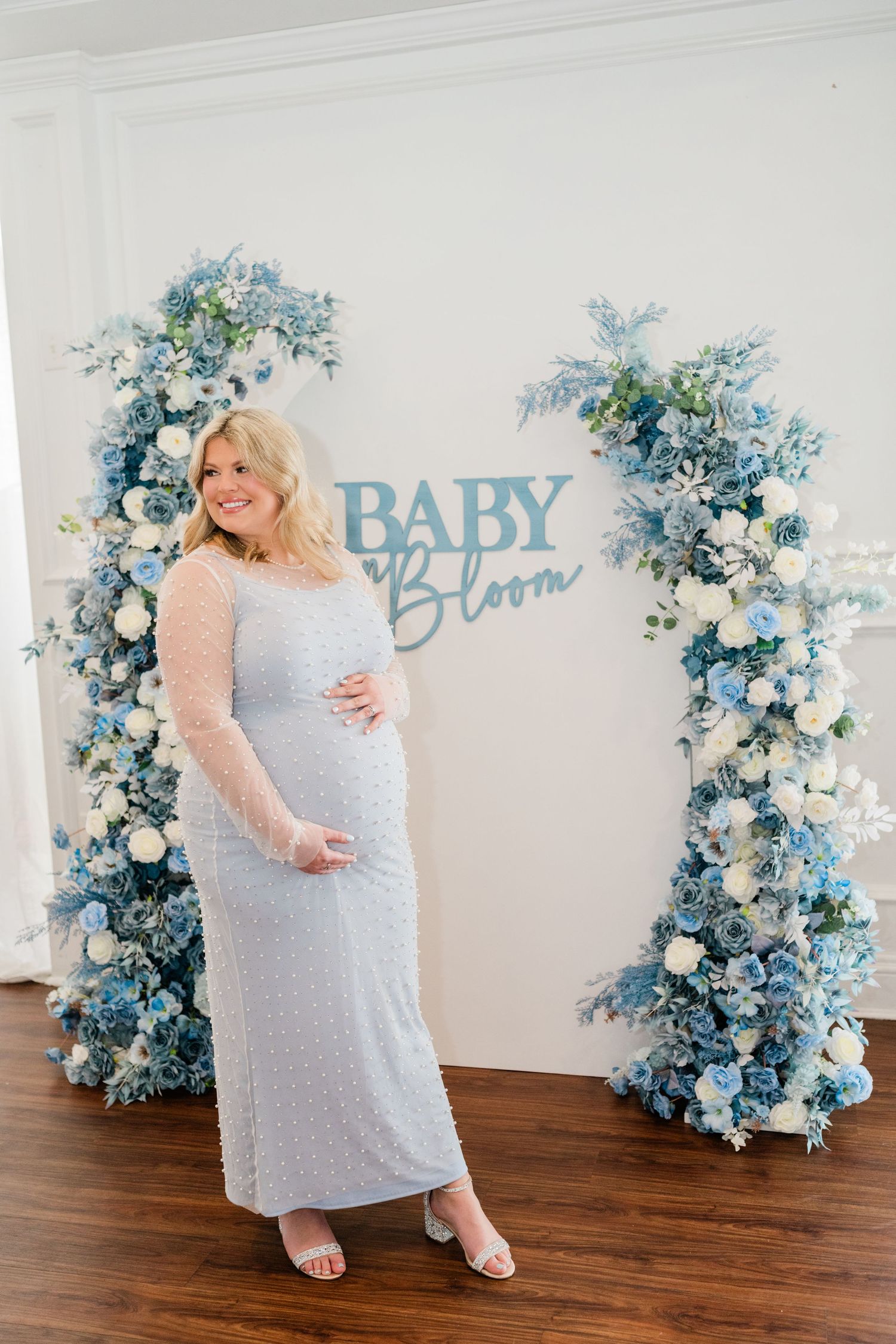 Expectant mother in white dress poses by blue floral arch with 'Baby Bloom' sign.