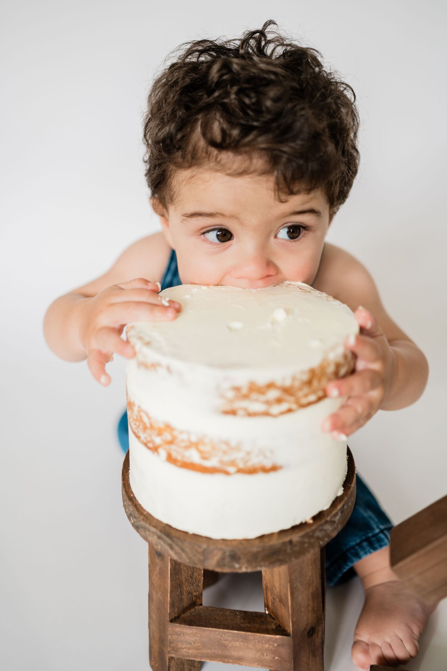Curly-haired toddler boy eating white frosted cake on wooden stool during first birthday cake smash photo session.
