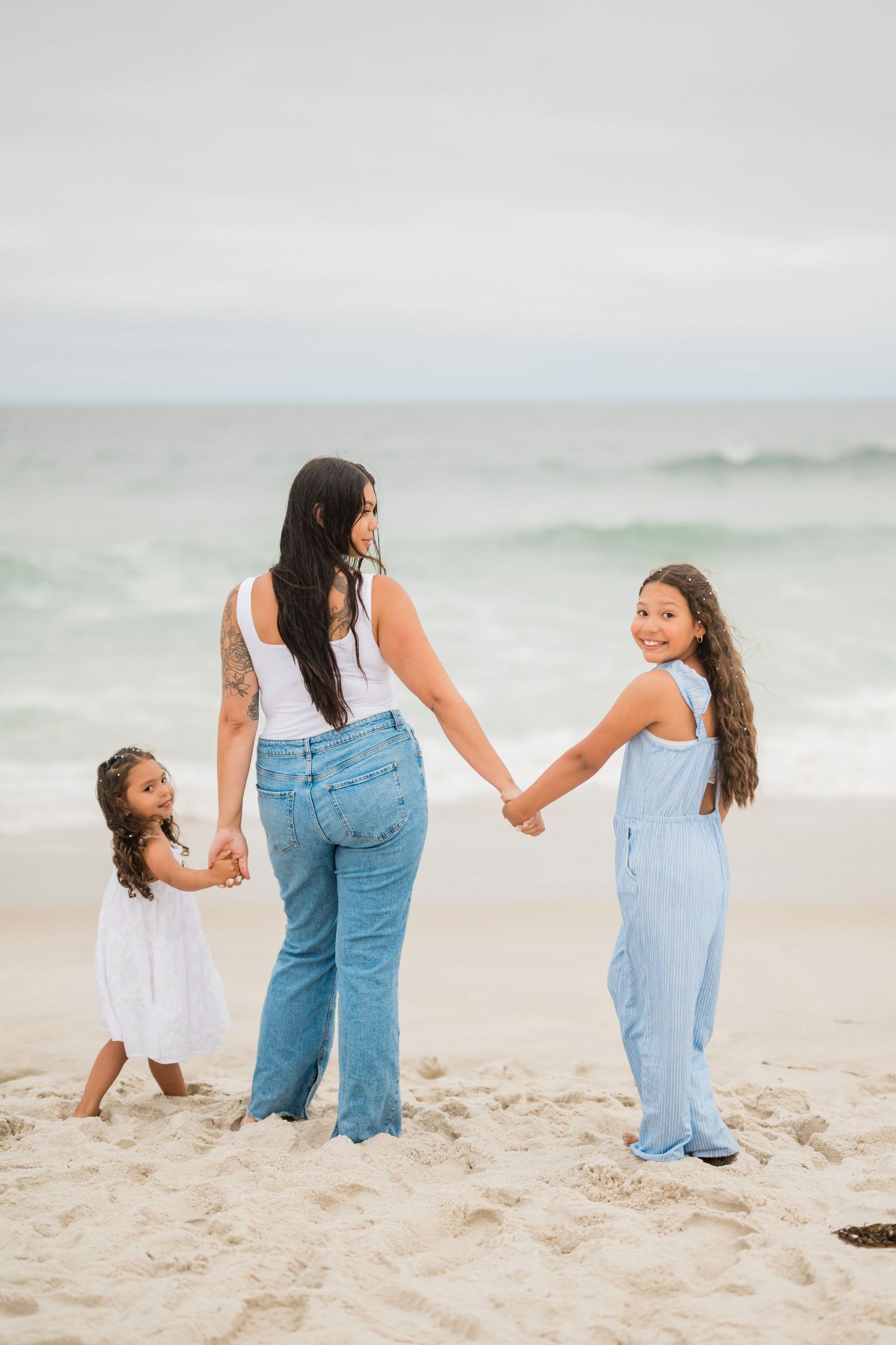A mother and two daughters hold hands on a sandy beach, with the older girl smiling back at the camera near ocean waves.