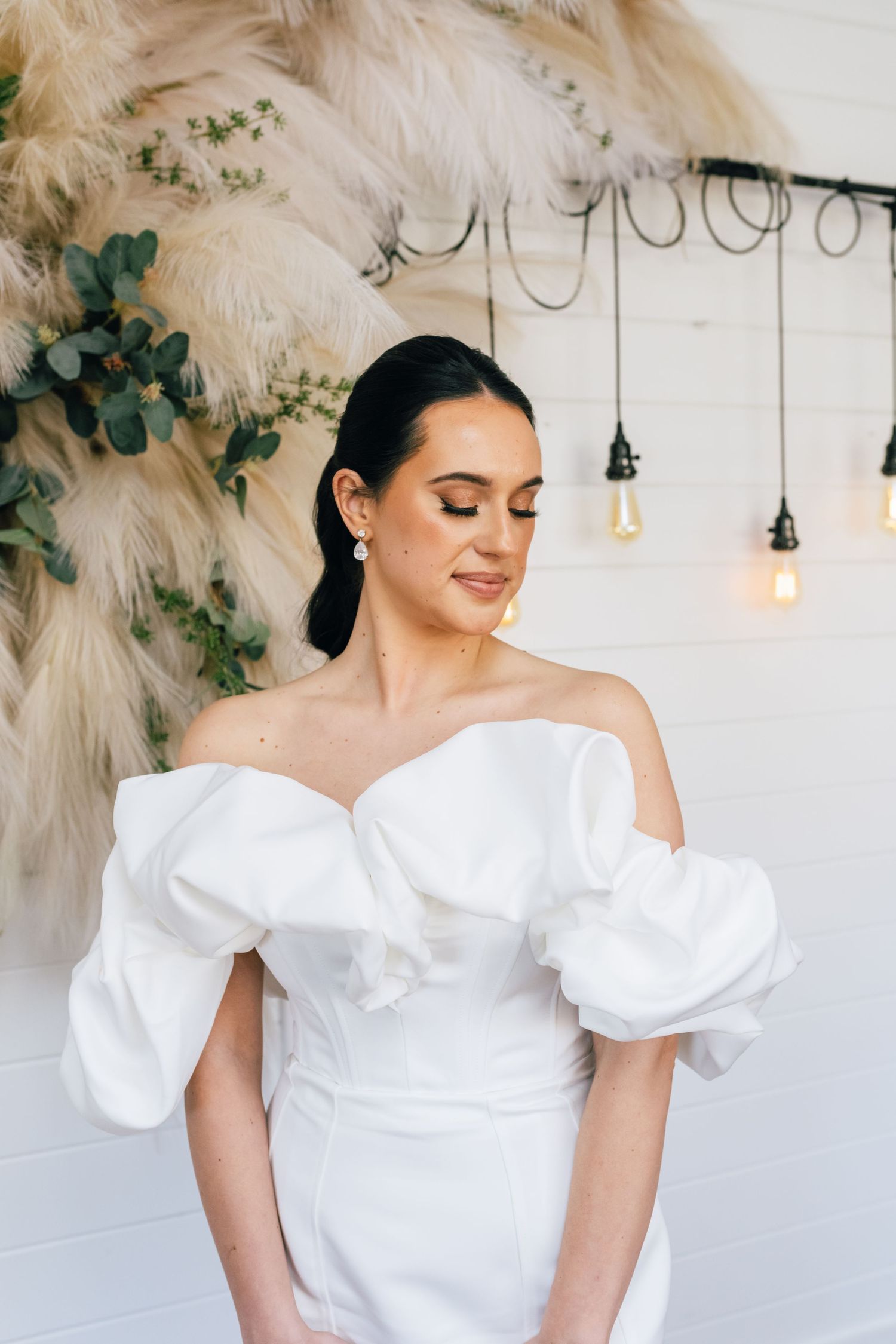 A person wearing an off-shoulder white gown poses against a backdrop with pampas grass and hanging lights.
