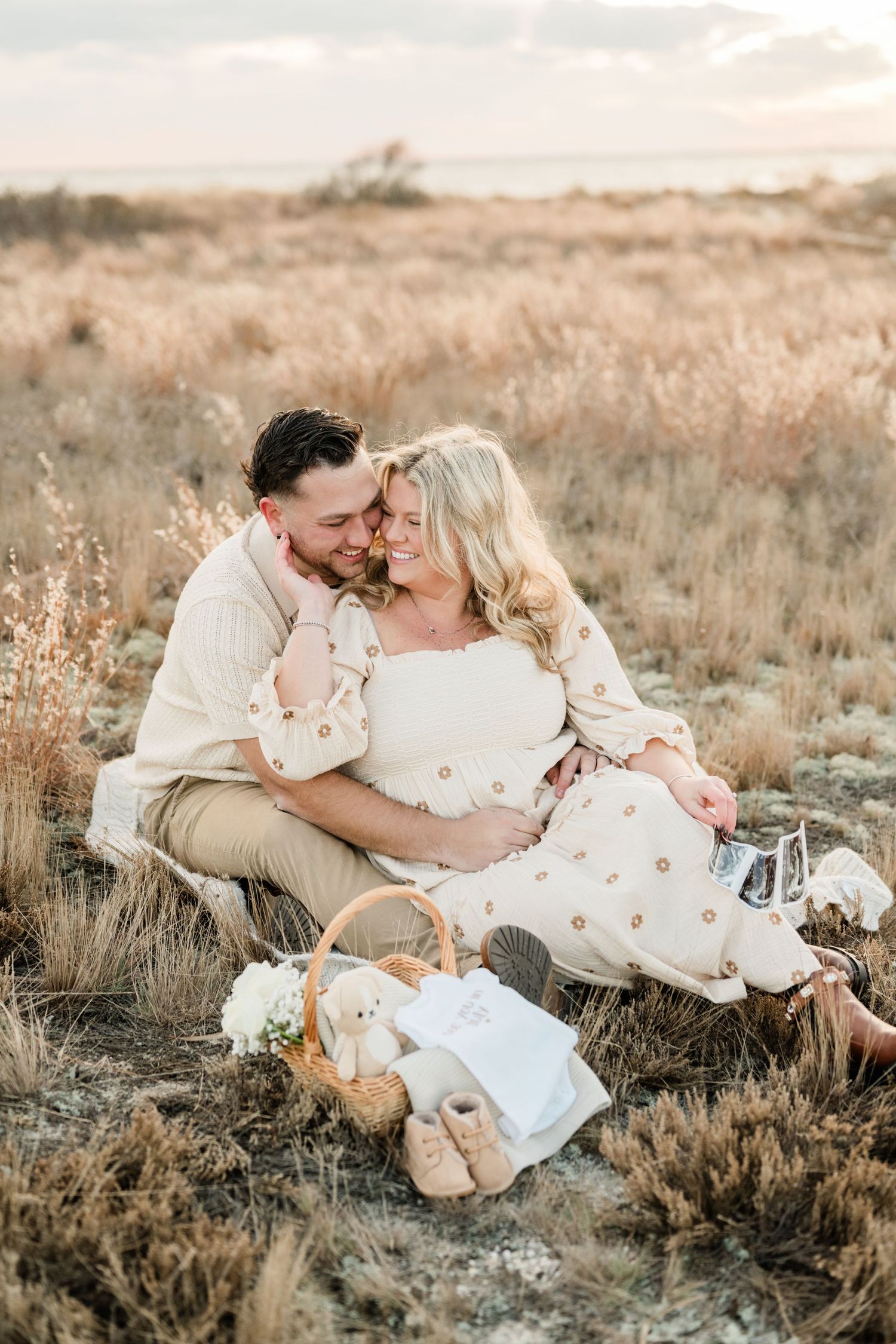 Couple sits in golden field at sunset with picnic basket and white dress.