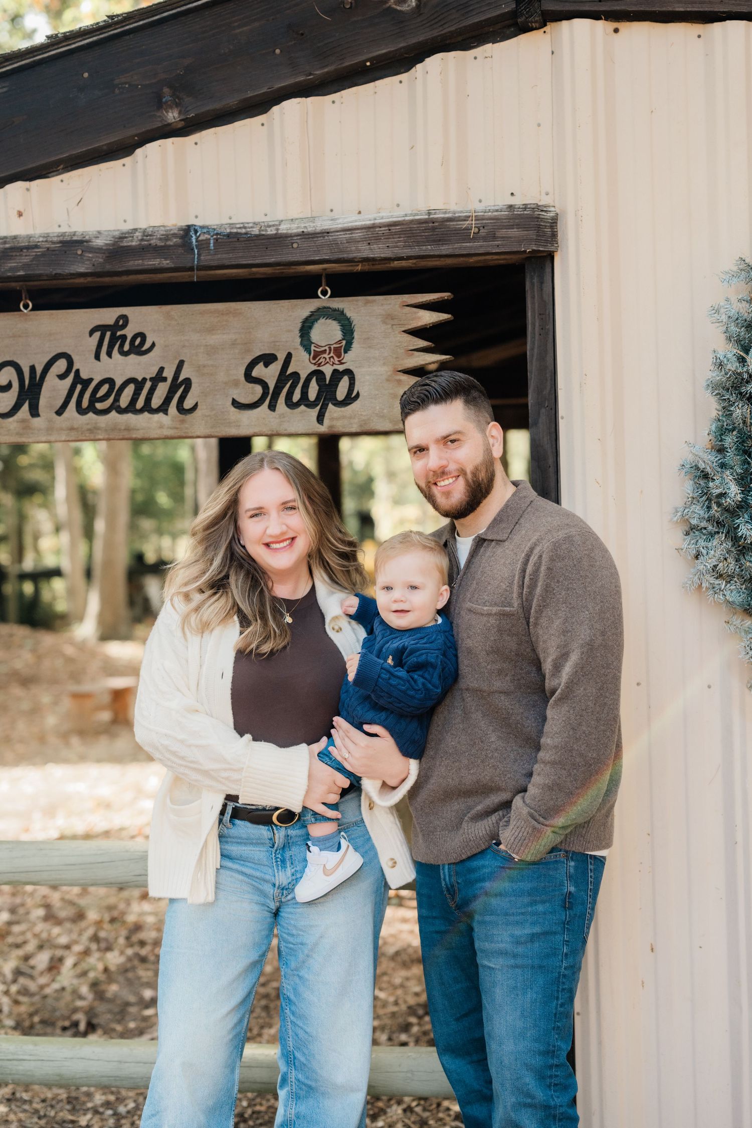 A smiling couple holds their baby boy outside The Wreath Shop, dressed in casual fall attire.