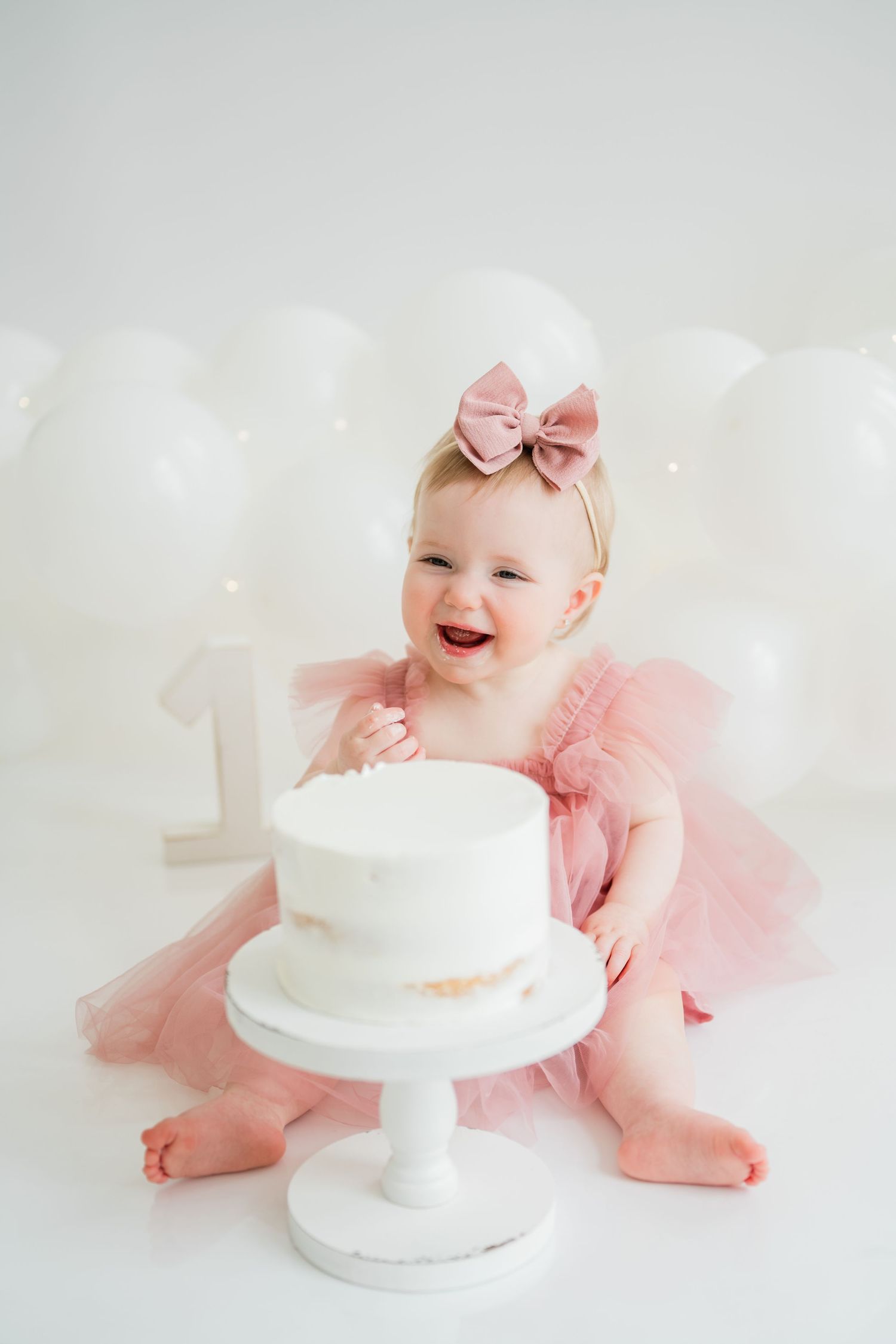 A joyful baby girl in a pink tutu dress celebrates her first birthday with a white cake and balloon decorations.