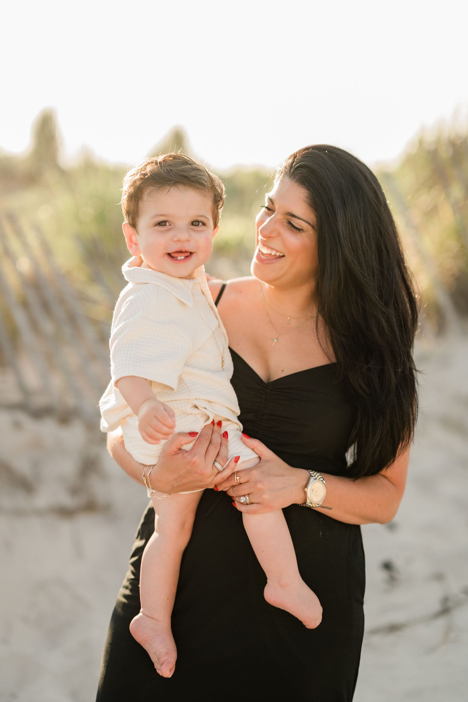 A smiling woman in a black dress holds a happy toddler boy on a sunny beach with sand dunes.