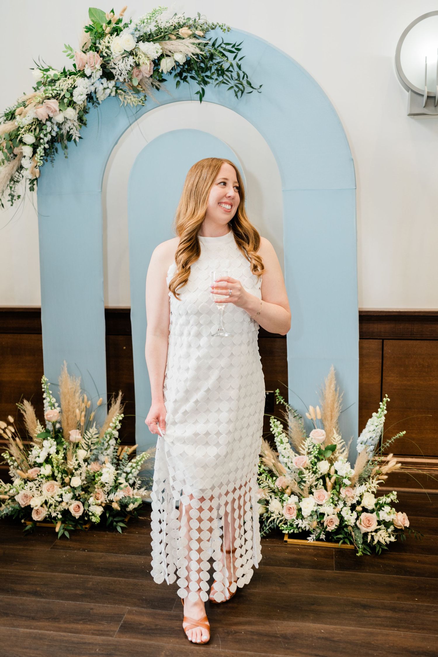 Person in white textured dress standing against blue arched backdrop with flowers.
