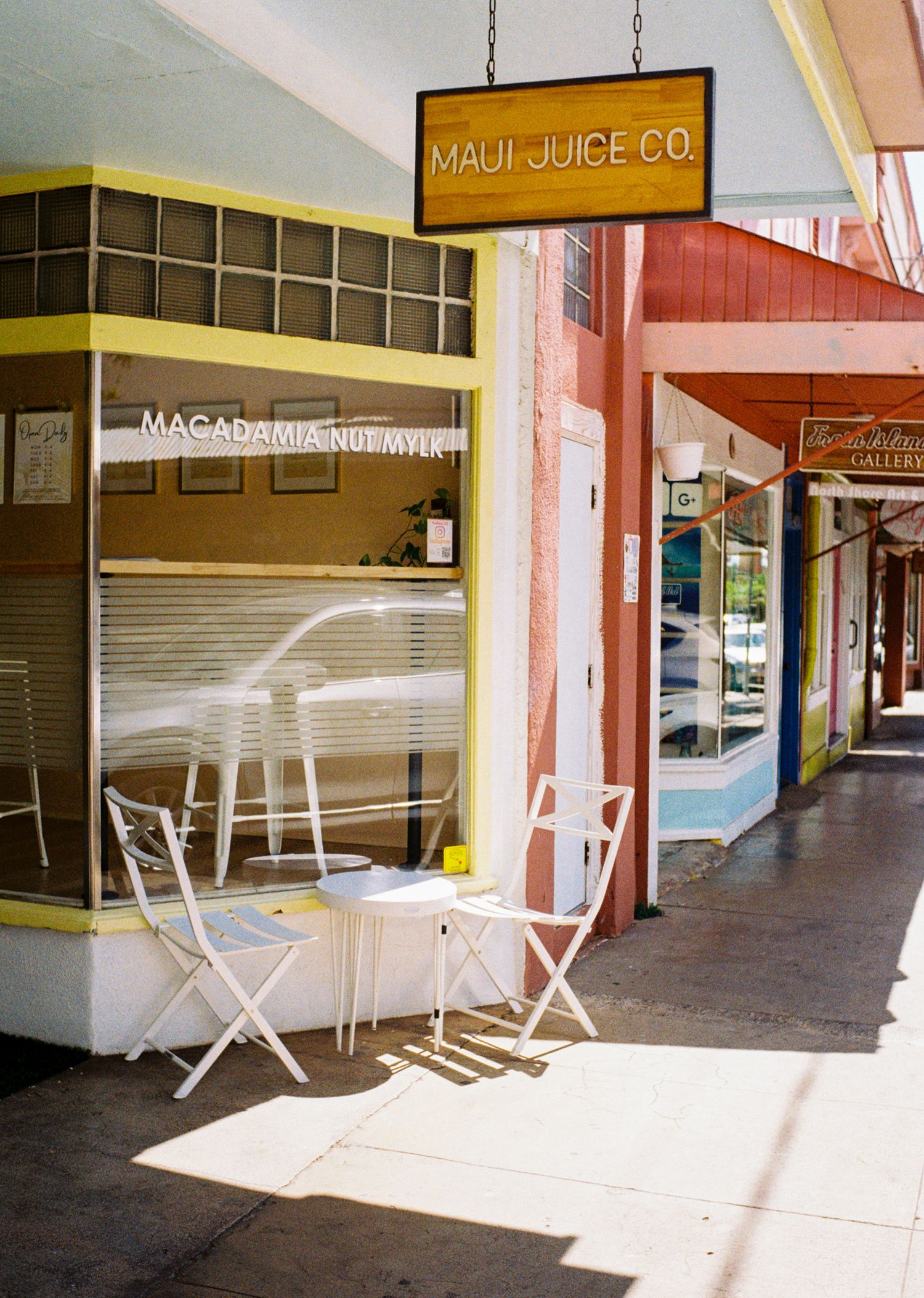 Empty storefront of Maui Juice Co with white chairs and tables on a sunny sidewalk.