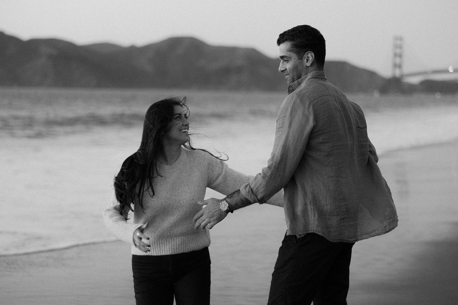A couple shares an intimate moment while walking together on a foggy beach near the Golden Gate Bridge.