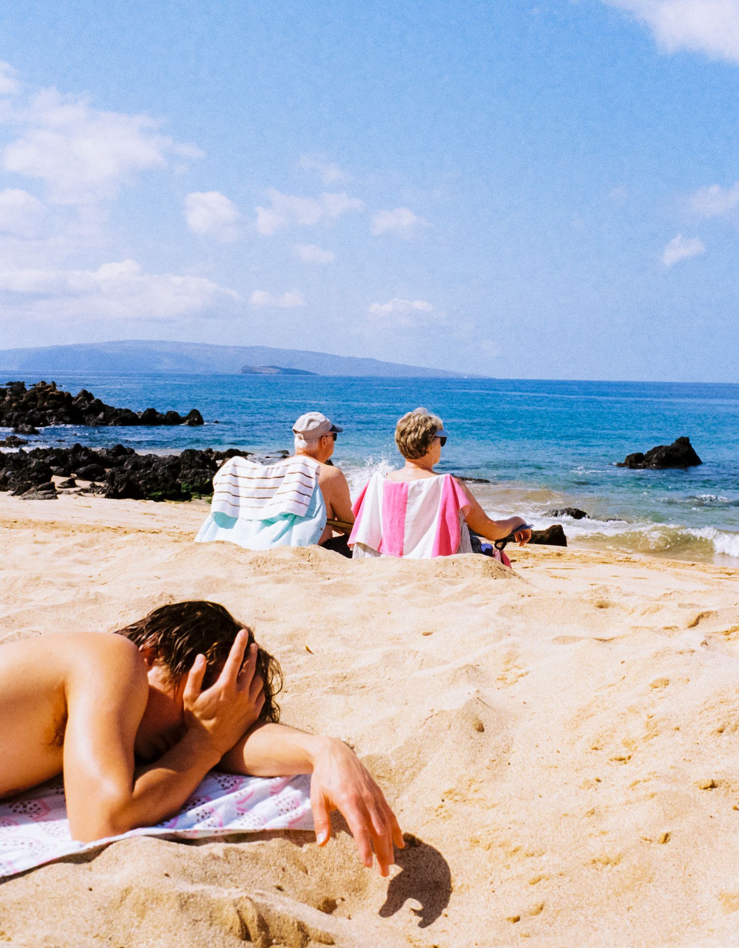 Beachgoers relaxing on sandy shore with blue ocean and volcanic rocks in background.