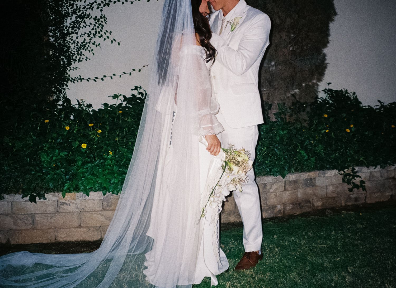 An elegant outdoor wedding moment with flowing white dress and veil against greenery.