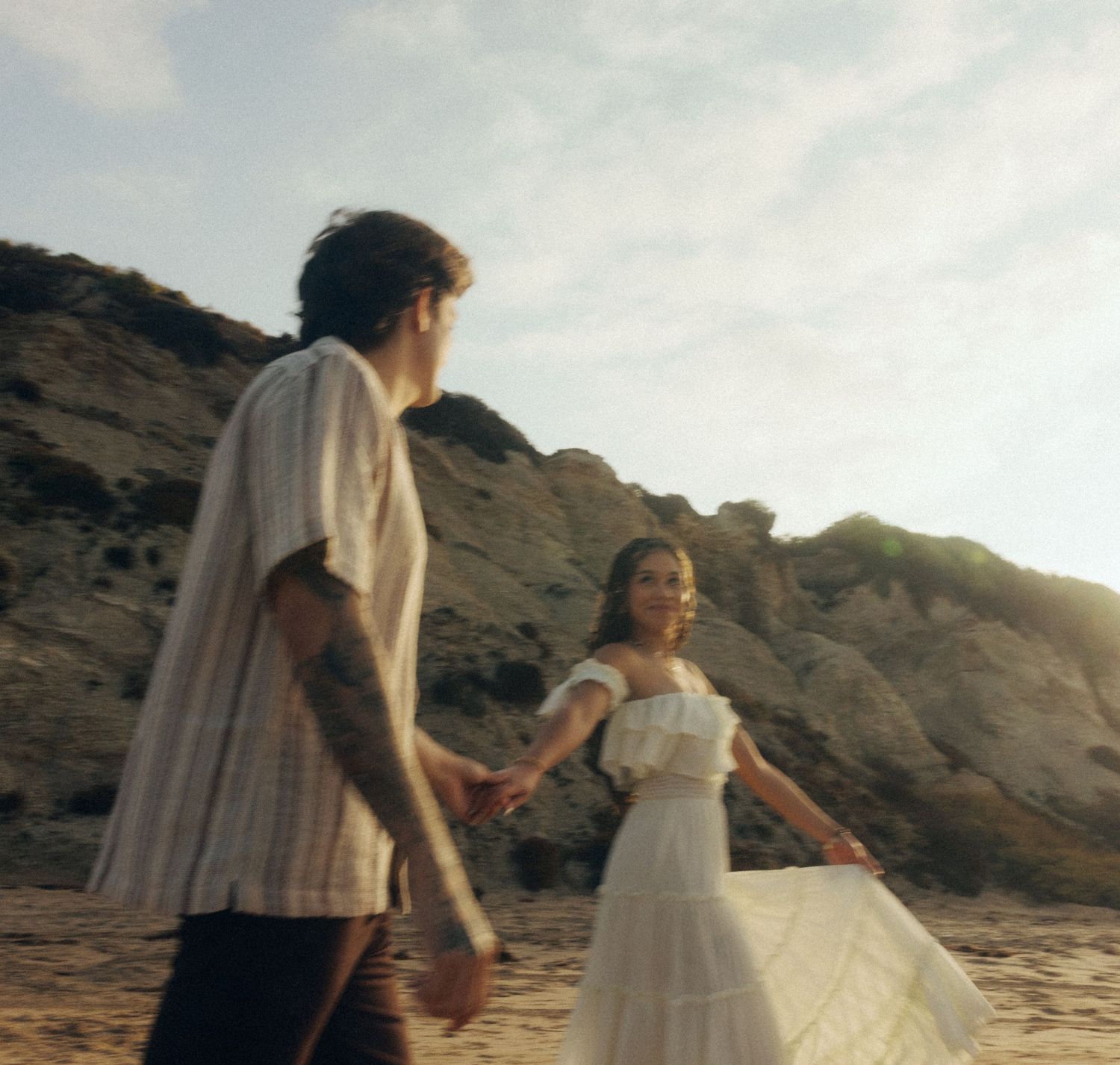 A couple in romantic beach scenery at sunset with the person in white dress twirling by the rocky cliffs.