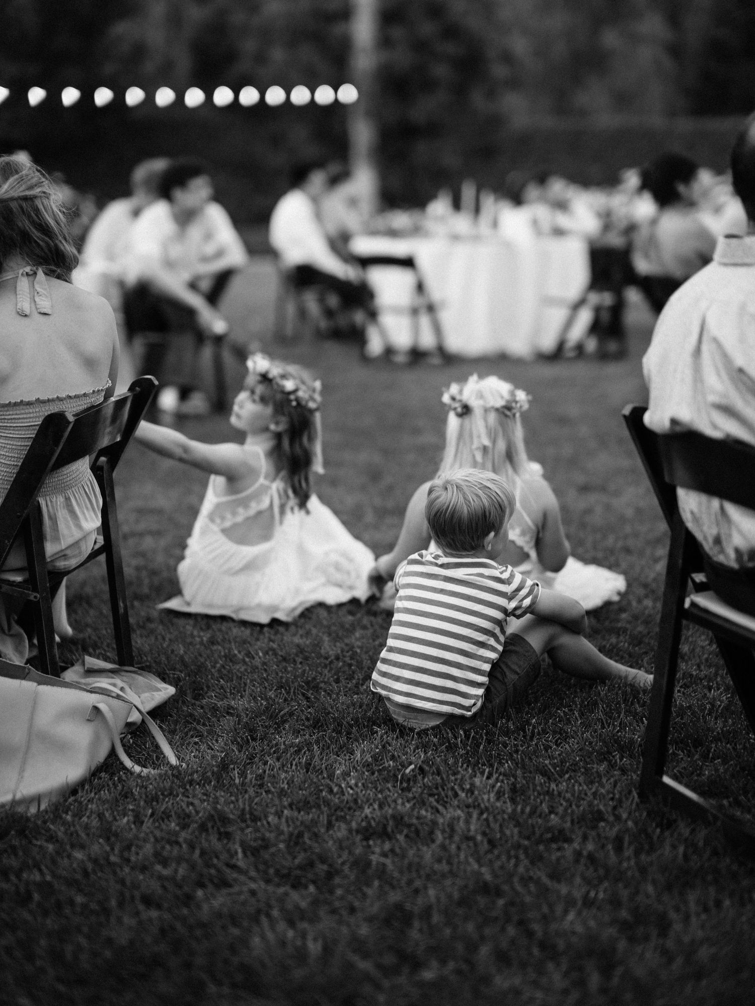 Children sit on grass watching an outdoor wedding reception with string lights and guests seated at tables.