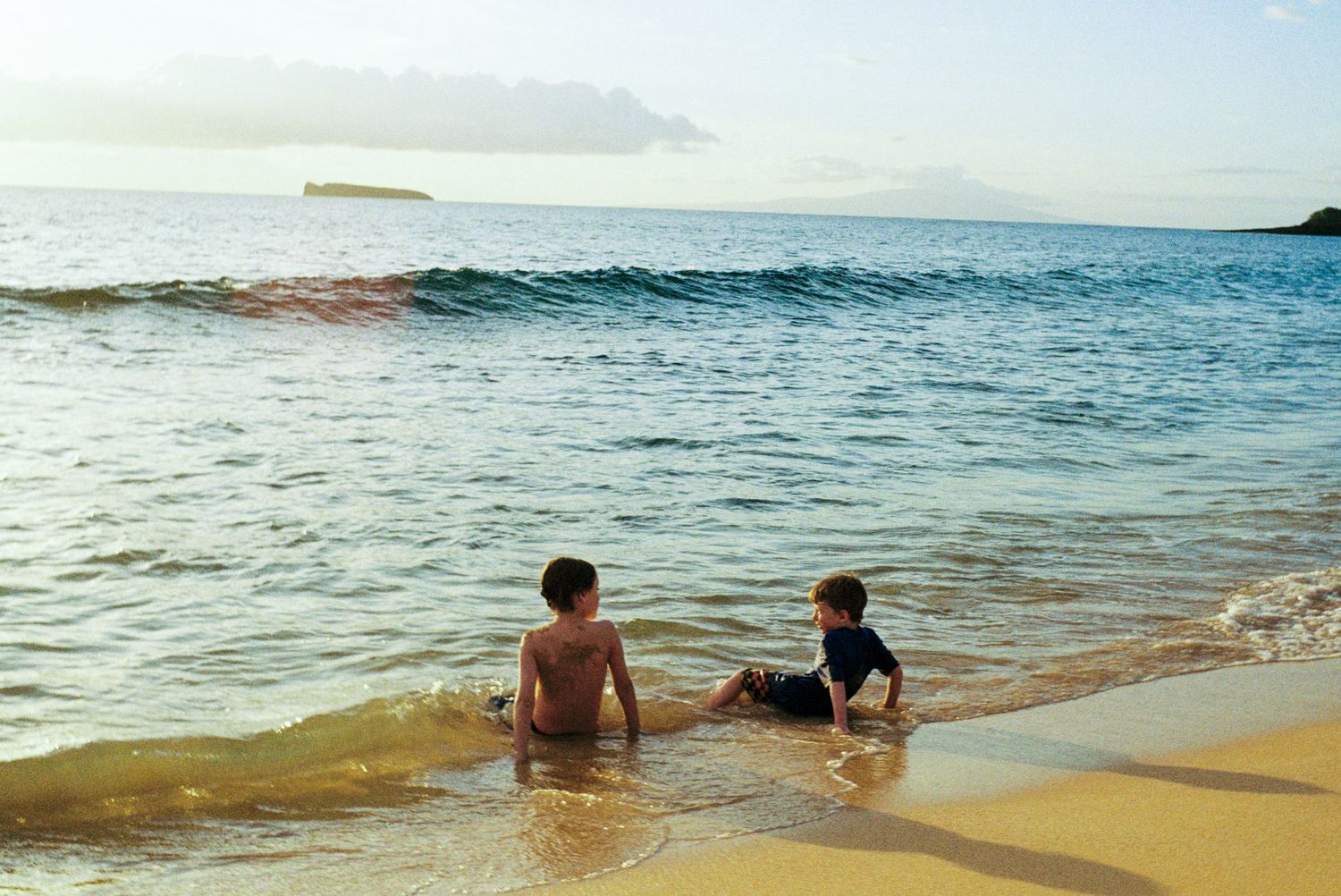 Two people relaxing in shallow ocean waves at sunset on a sandy beach.