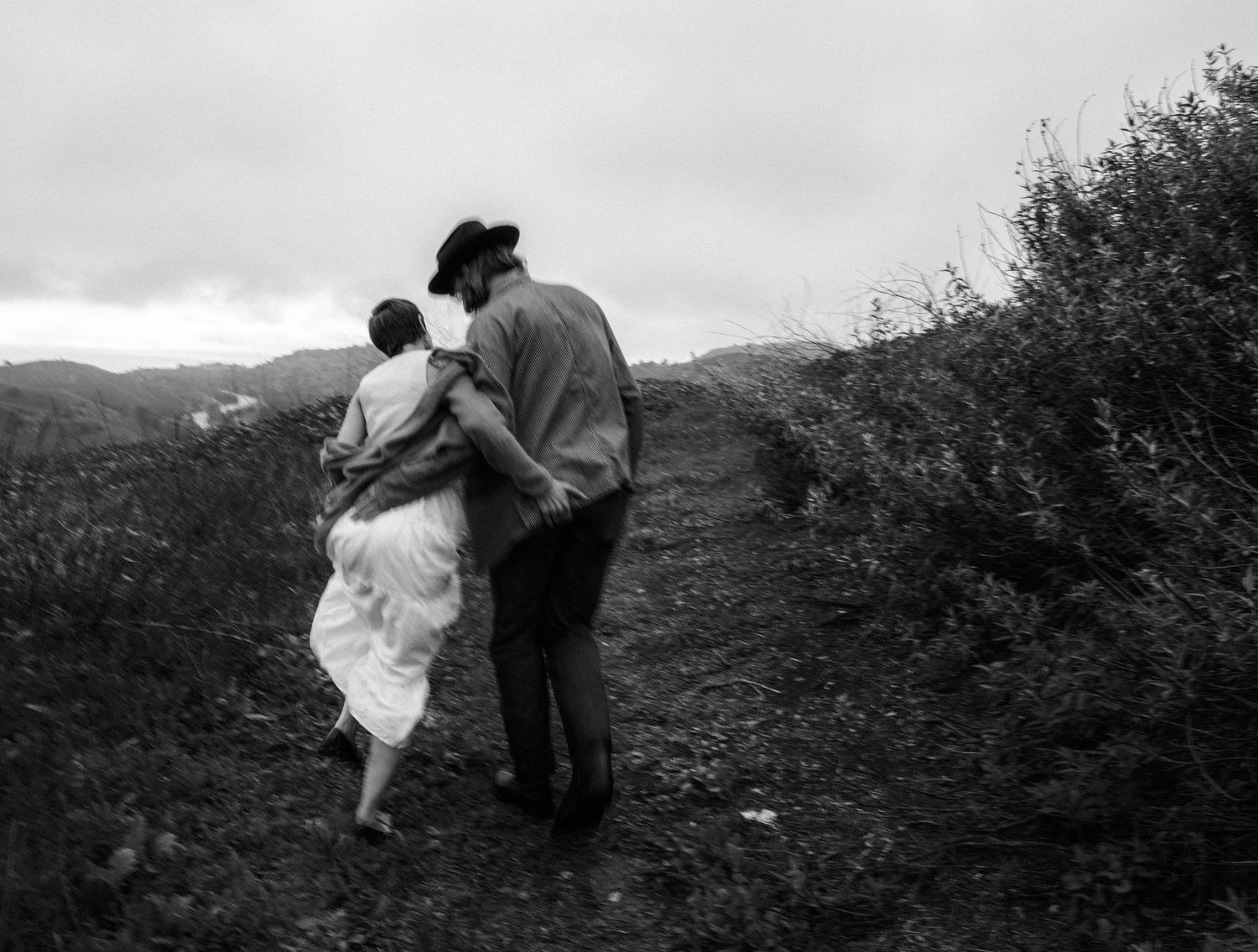 Two figures embrace while walking along a hilltop path against a cloudy mountain landscape.