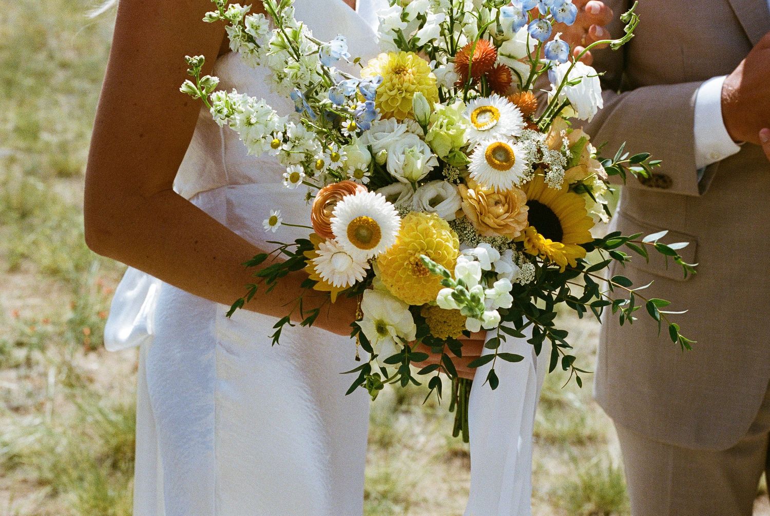 A rustic bridal bouquet featuring bright sunflowers, white daisies and delicate greenery against a white wedding dress.