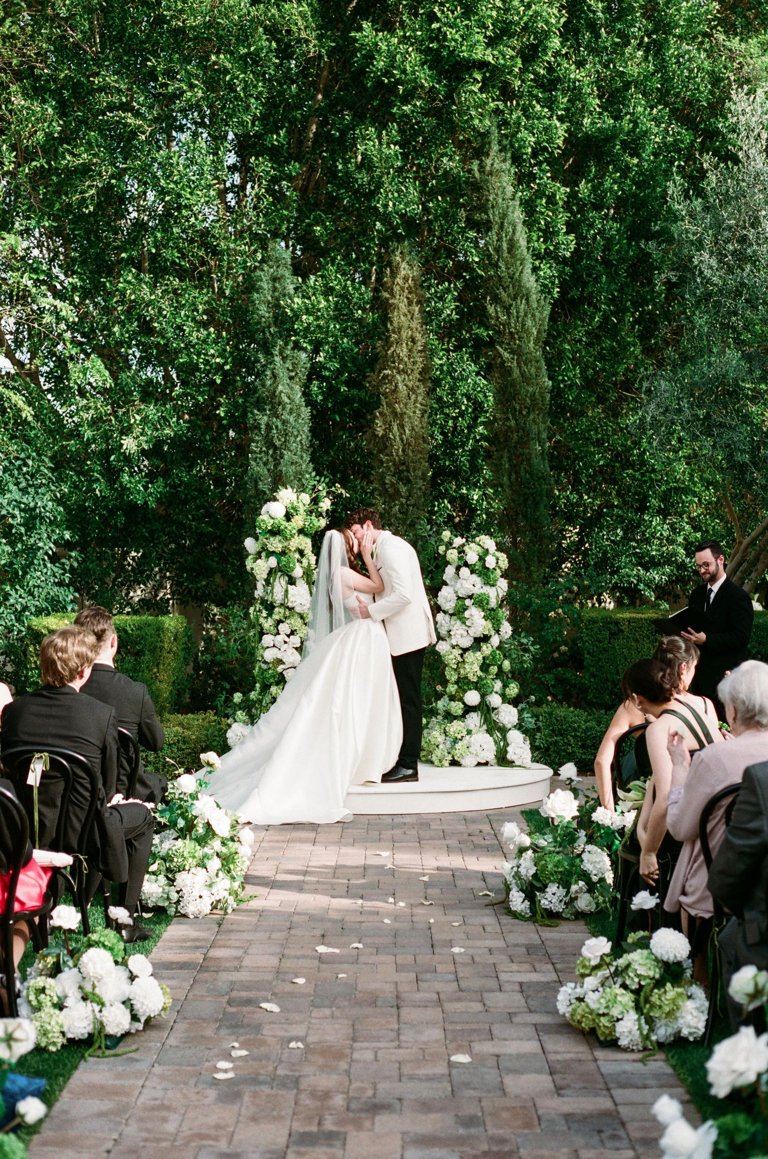 White floral arrangements line a brick pathway at an outdoor wedding ceremony in a lush garden setting.
