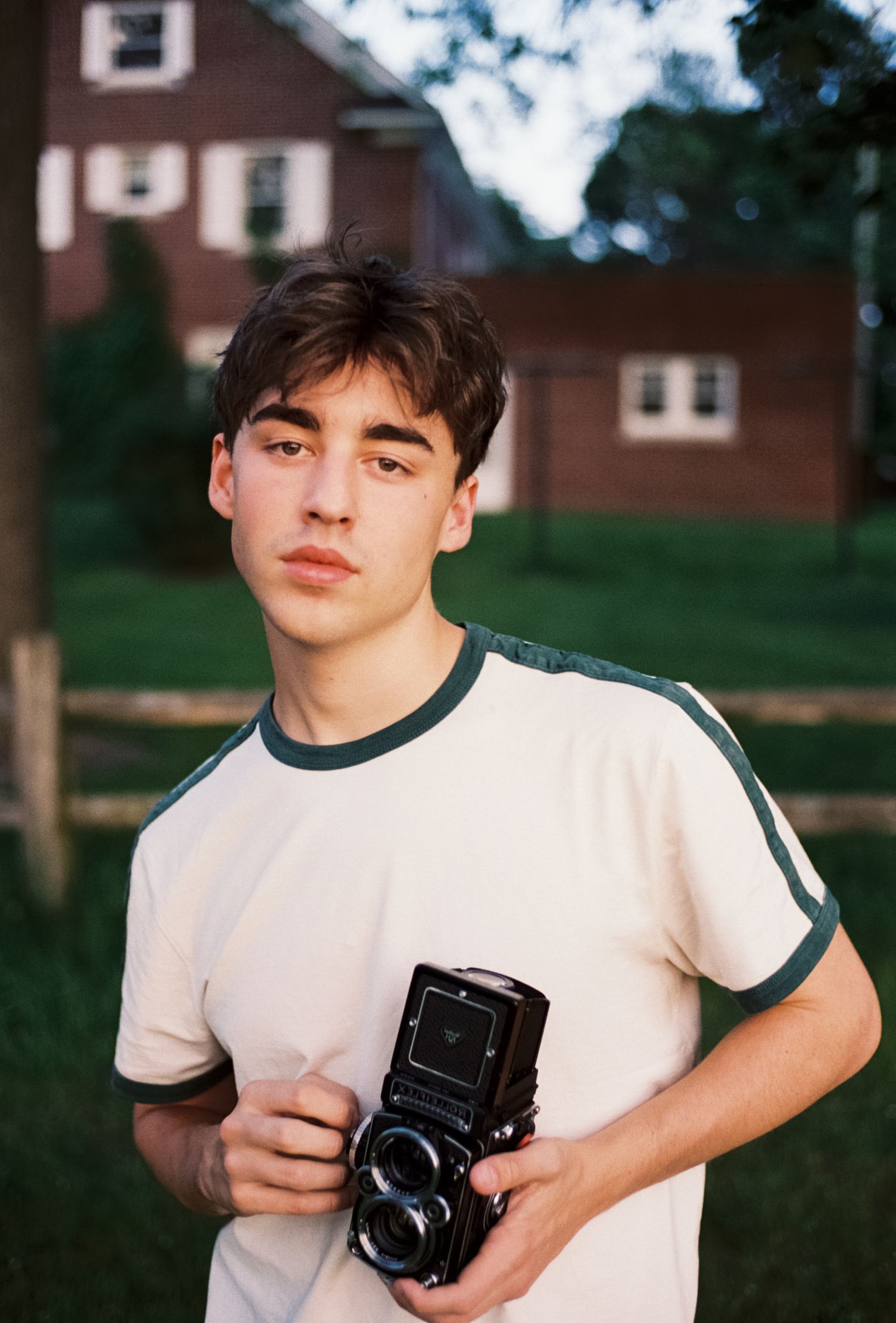 A person in a white ringer t-shirt holds a vintage twin-lens reflex camera outdoors with brick buildings in the background.