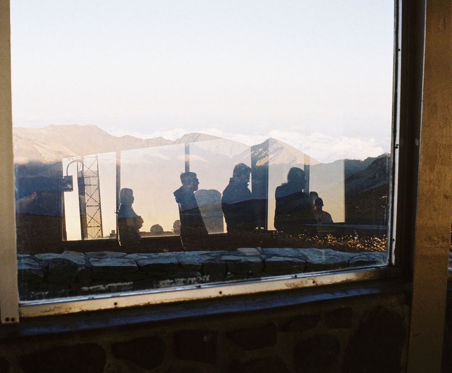 Silhouettes of observers admiring mountain view through large window at sunset.