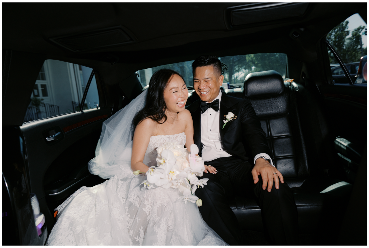 A couple sits together in the back of a luxury car, with a white wedding dress and formal tuxedo.