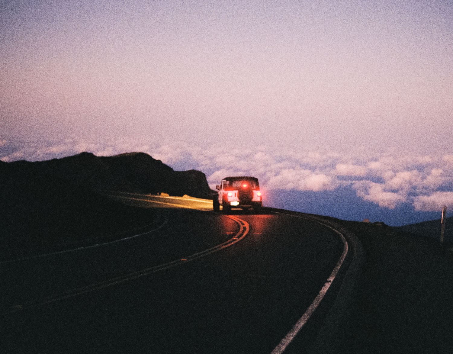 Car headlights illuminate a winding mountain road above clouds at dusk.