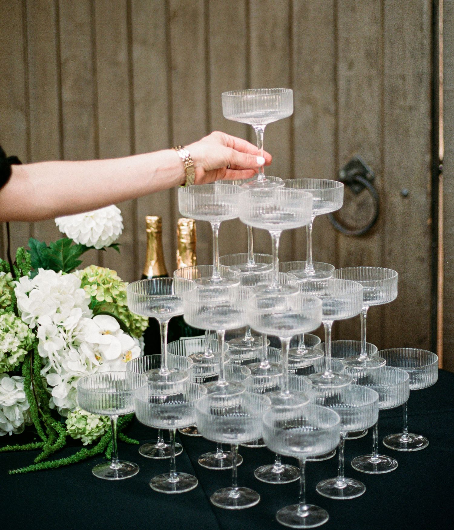 A tiered display of elegant champagne coupes arranged in rows on a rustic wooden surface with white floral decorations.