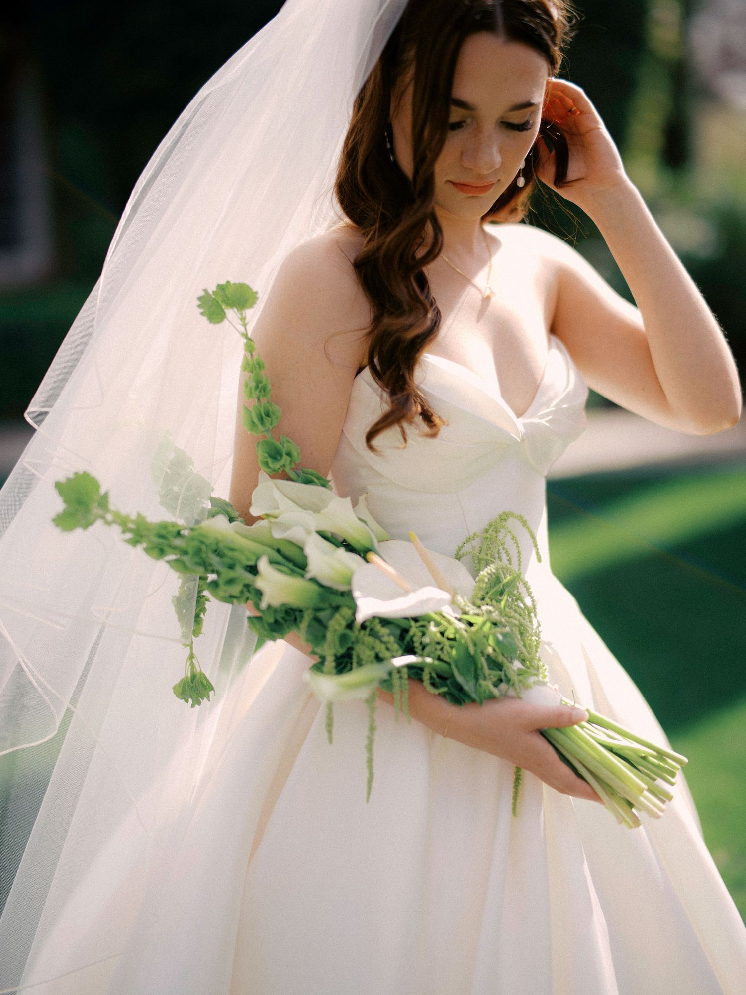 Bride in white strapless ballgown holding a natural greenery bouquet in outdoor garden setting.