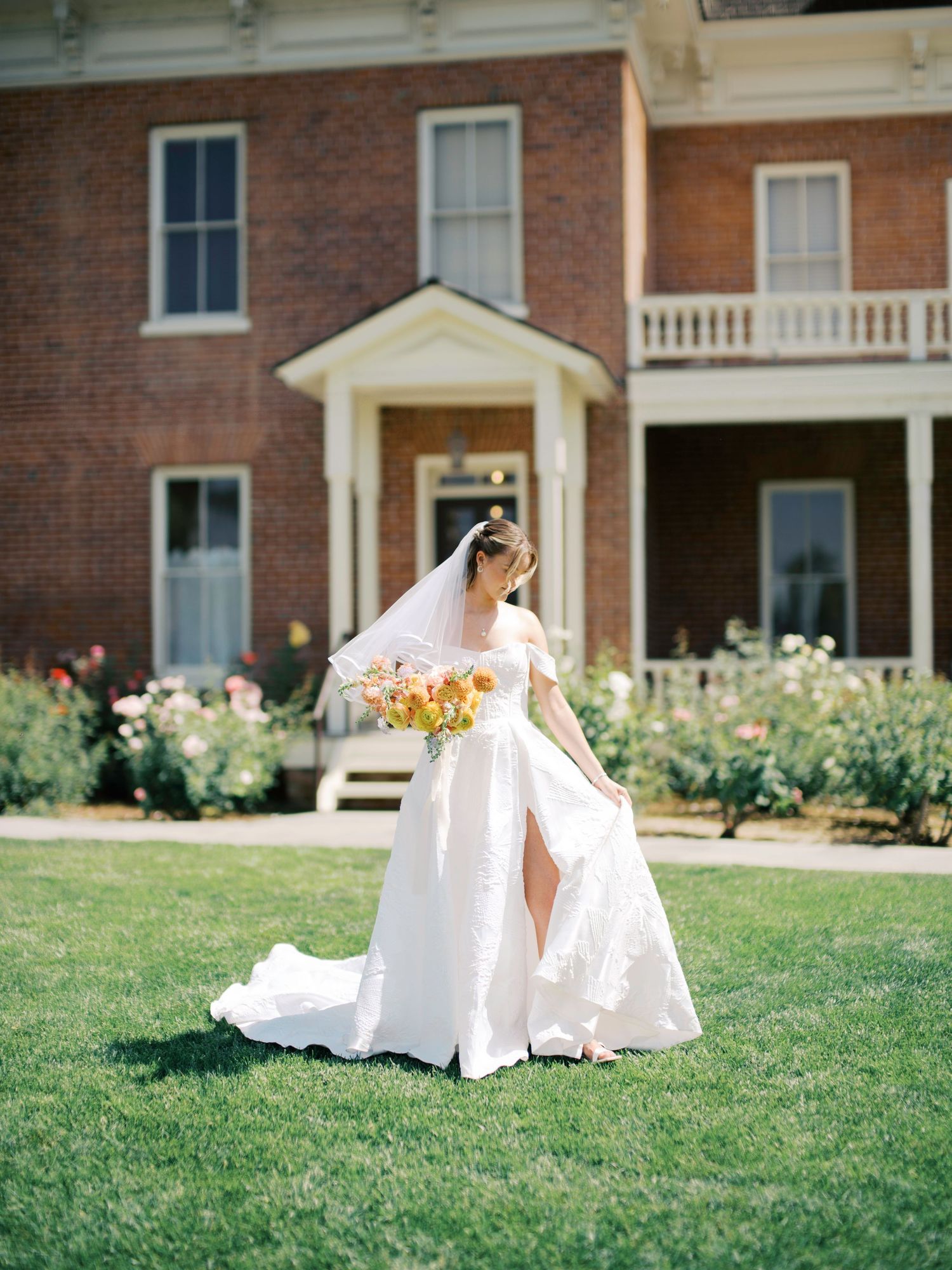 Beautiful bride in flowing white gown holds bouquet while standing on lawn in front of historic brick mansion.